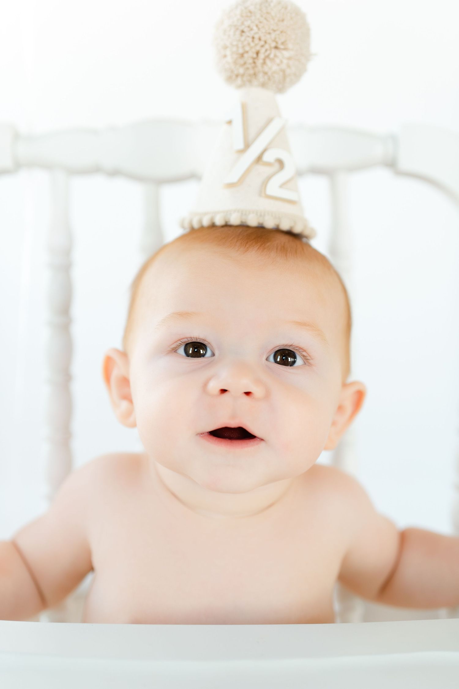 Adorable baby wearing a cream half birthday party hat with pom pom sits in a white high chair.