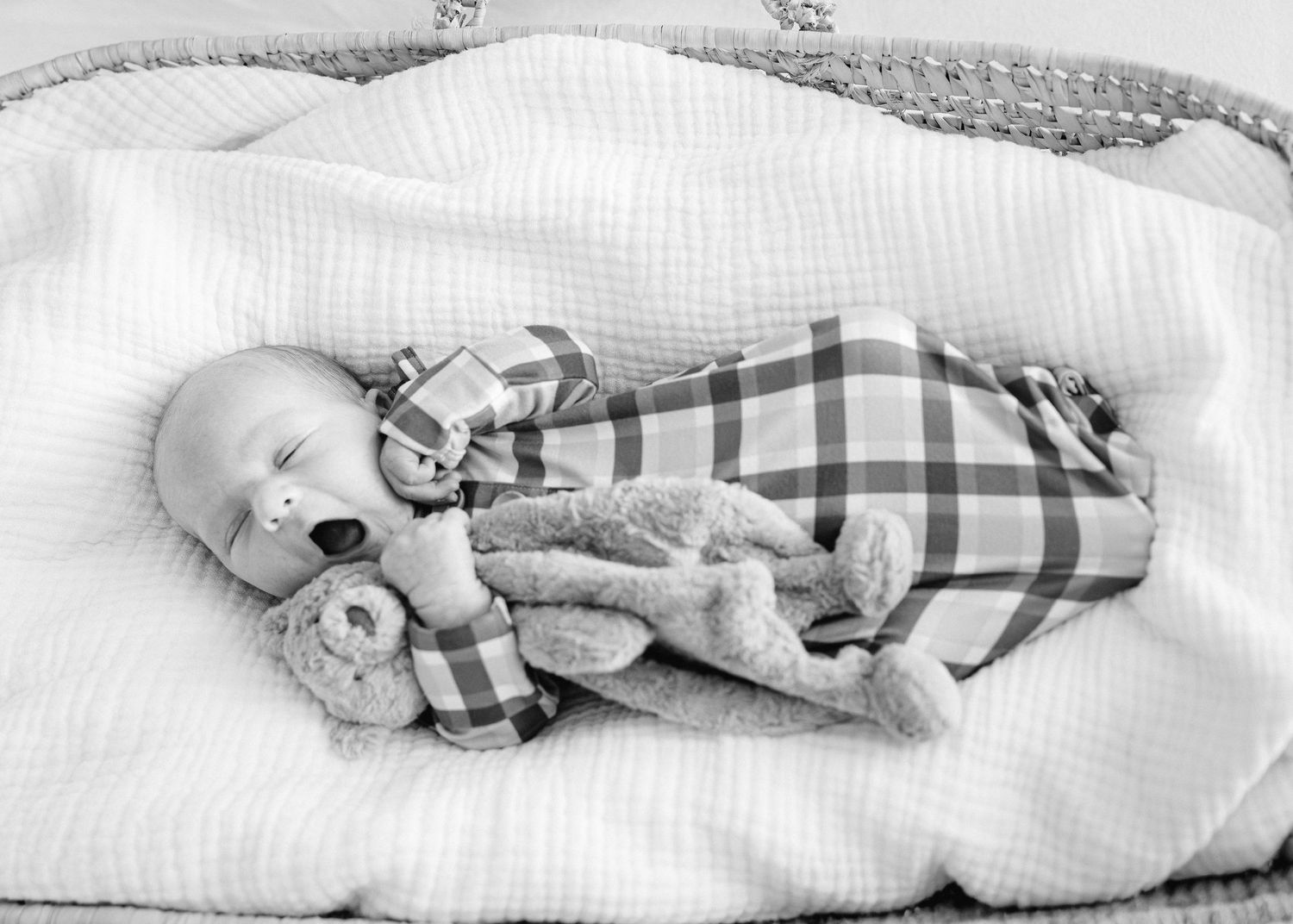 Sleeping newborn baby in plaid onesie cuddling a teddy bear in a white basket, black and white photo.