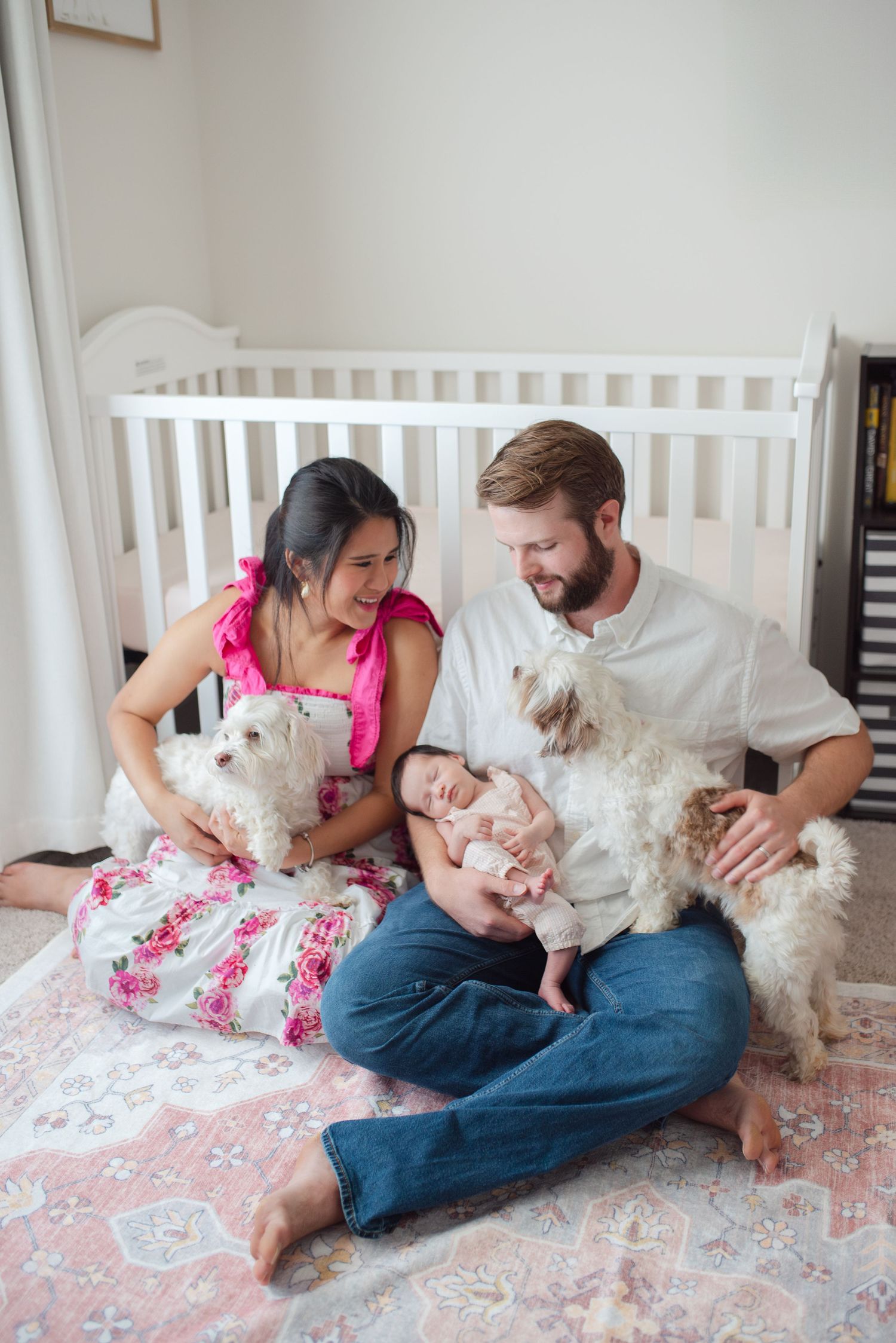 Smiling family with newborn and two white fluffy dogs sitting together near baby crib.