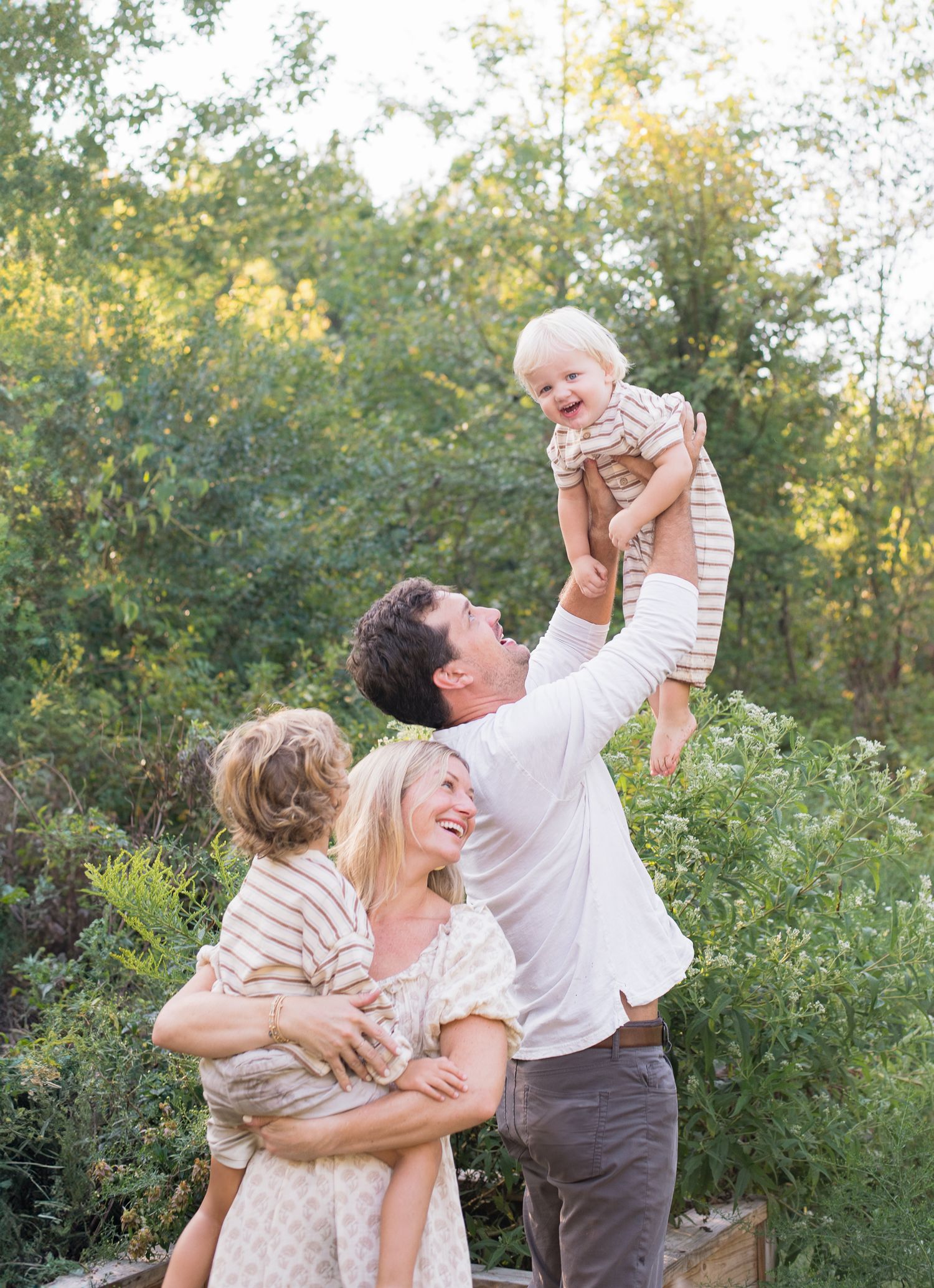 Playful family moment with father lifting laughing baby in the air while mother holds toddler outdoors.
