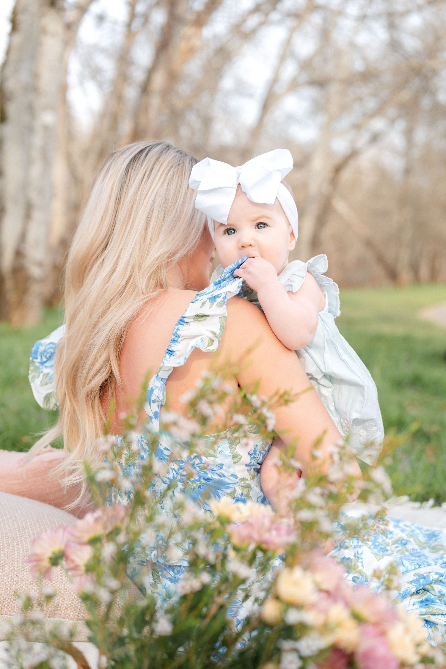 A blonde mother kisses her baby girl in a white bow headband while sitting among spring flowers.