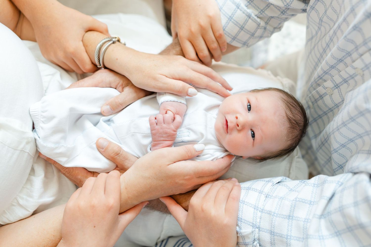 Newborn baby in white onesie being held by multiple family members hands, looking alert and calm.