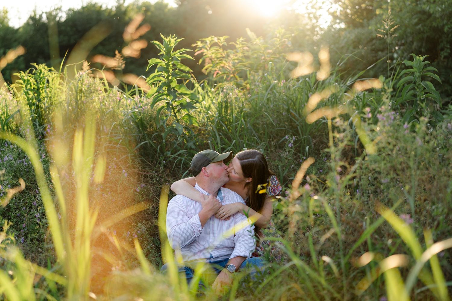 Couple sharing a romantic kiss while sitting in a sunlit meadow surrounded by tall green grass and wildflowers.