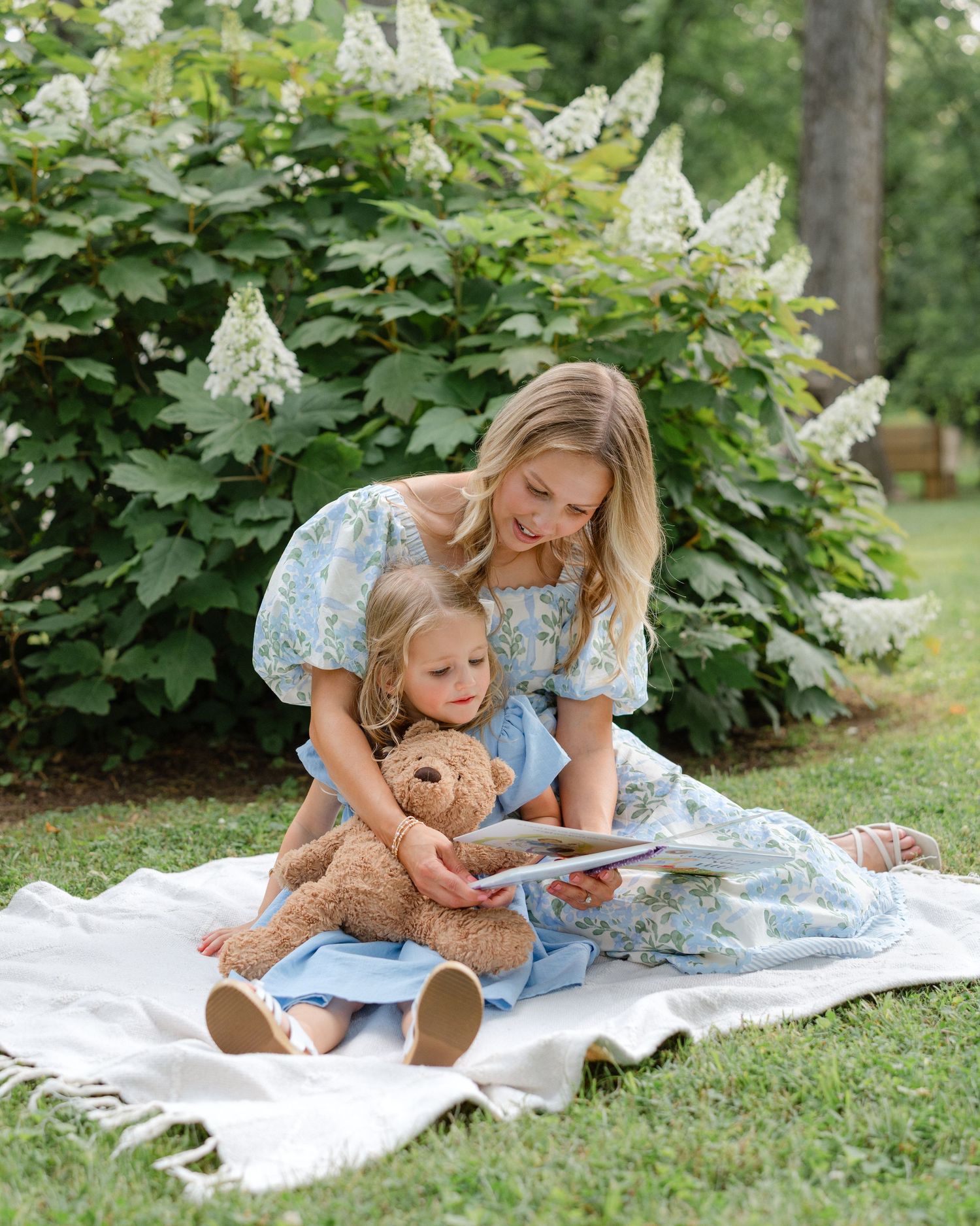 A mother and young daughter in matching blue floral dresses read a book together on a blanket outdoors.