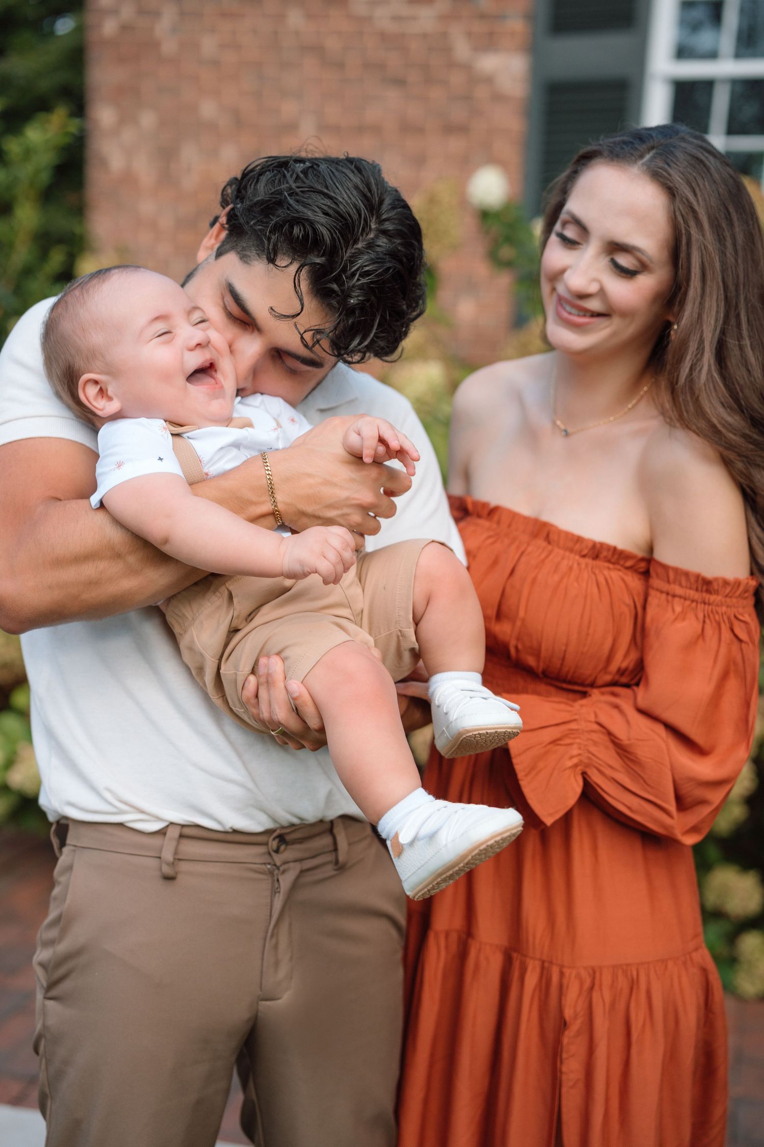 Young couple laughing with their baby boy outdoors near a brick building during a summer family photo session.