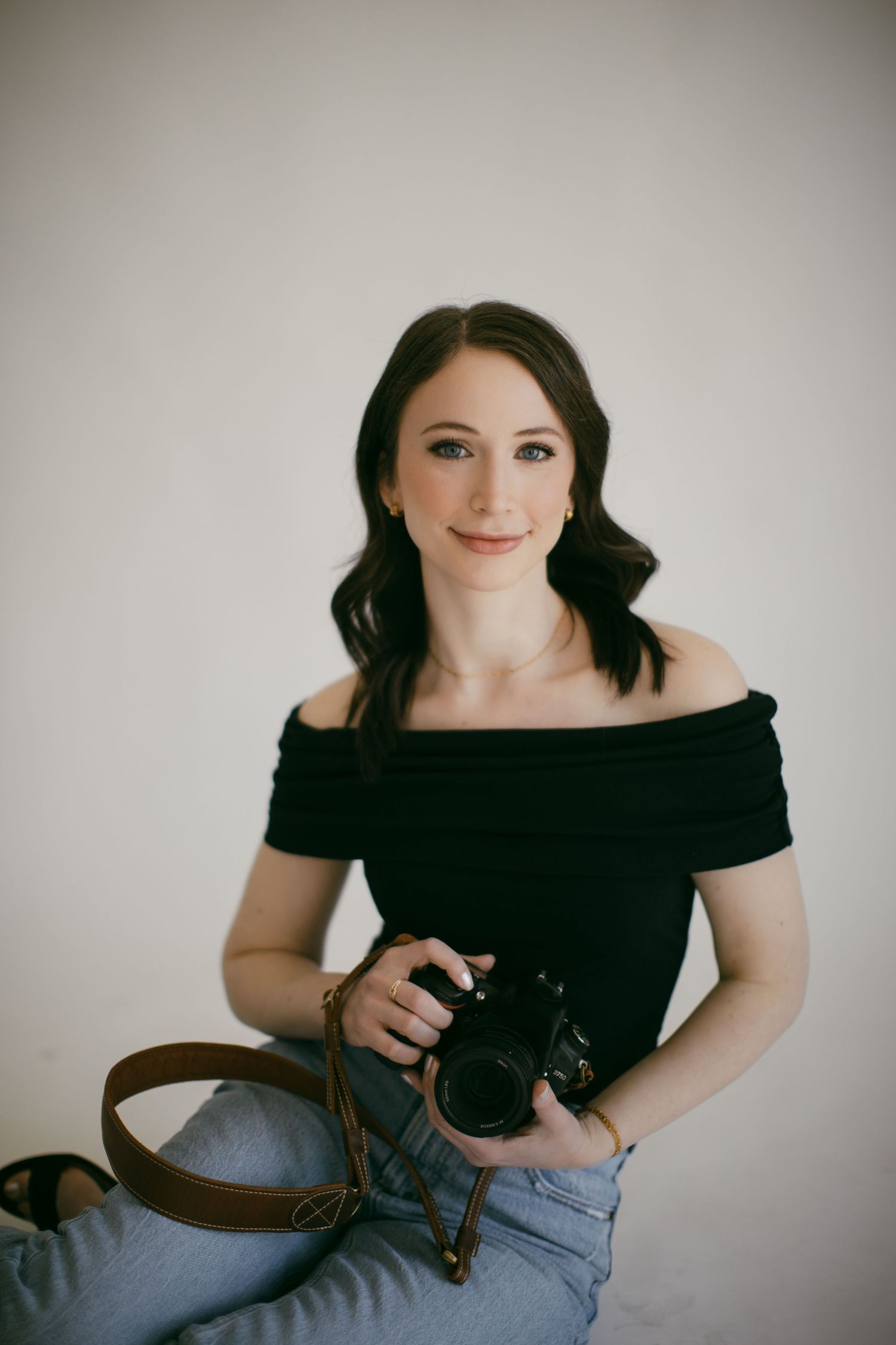 Professional female photographer in black off-shoulder top holding a camera while sitting against a white background.