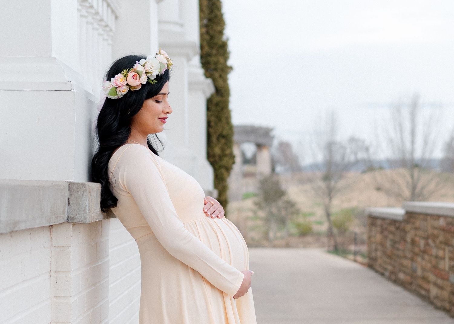 Pregnant woman in cream maternity gown and floral crown leans against white wall outdoors.