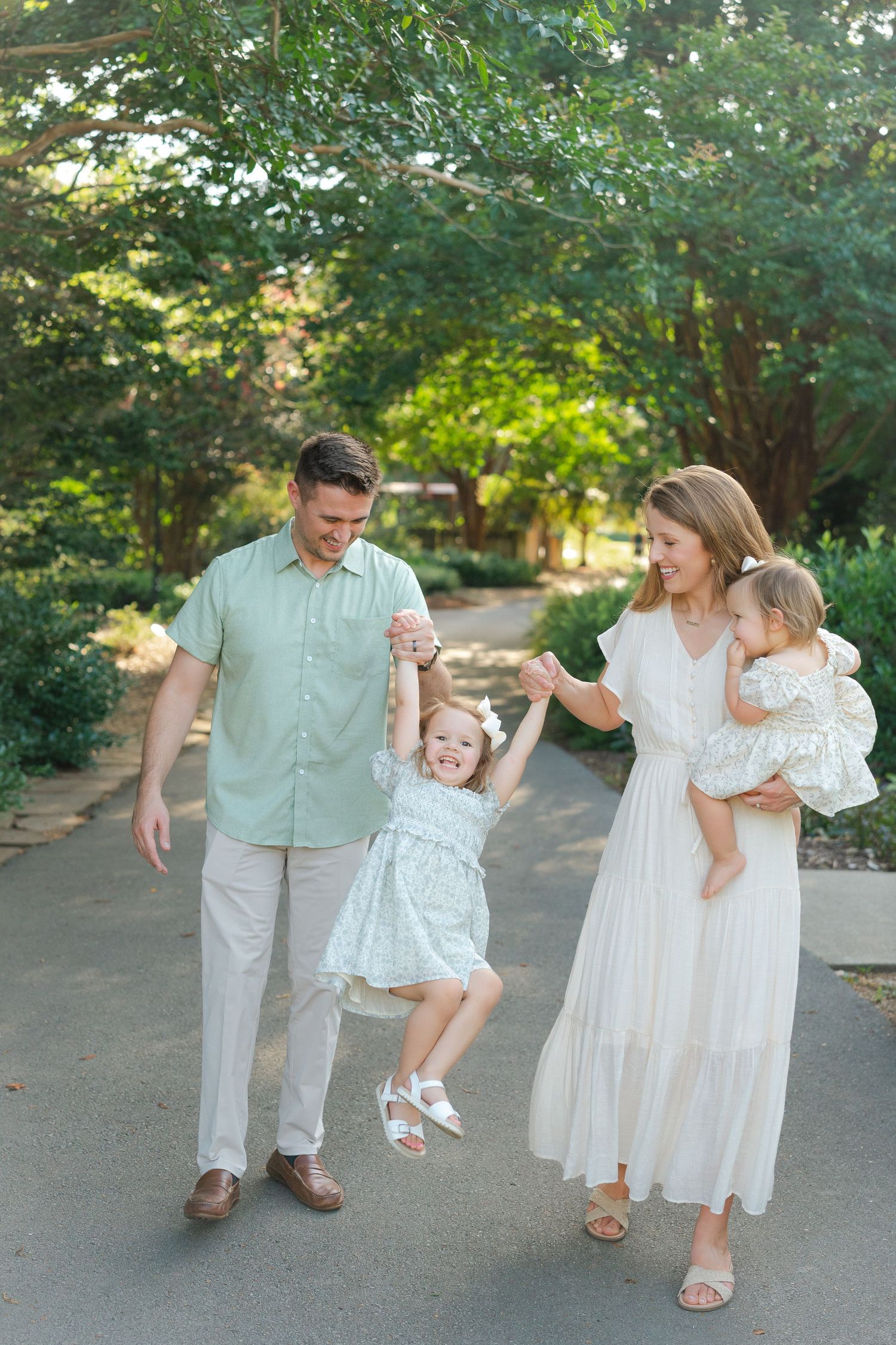 A joyful family swings their toddler daughter while walking in a lush green park, with mom holding a baby girl.