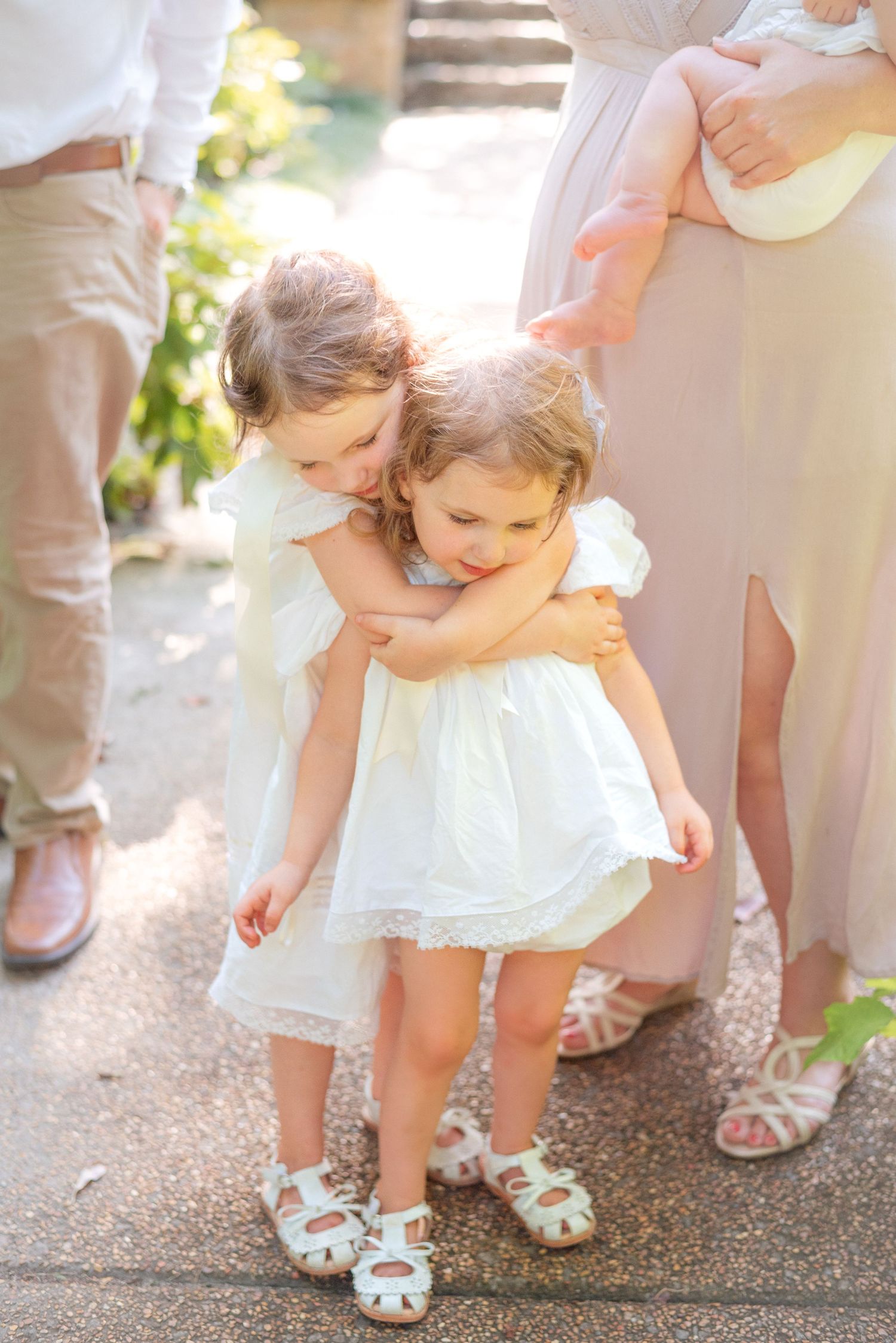Two young sisters in matching white dresses sharing a sweet hug outdoors in soft golden sunlight.