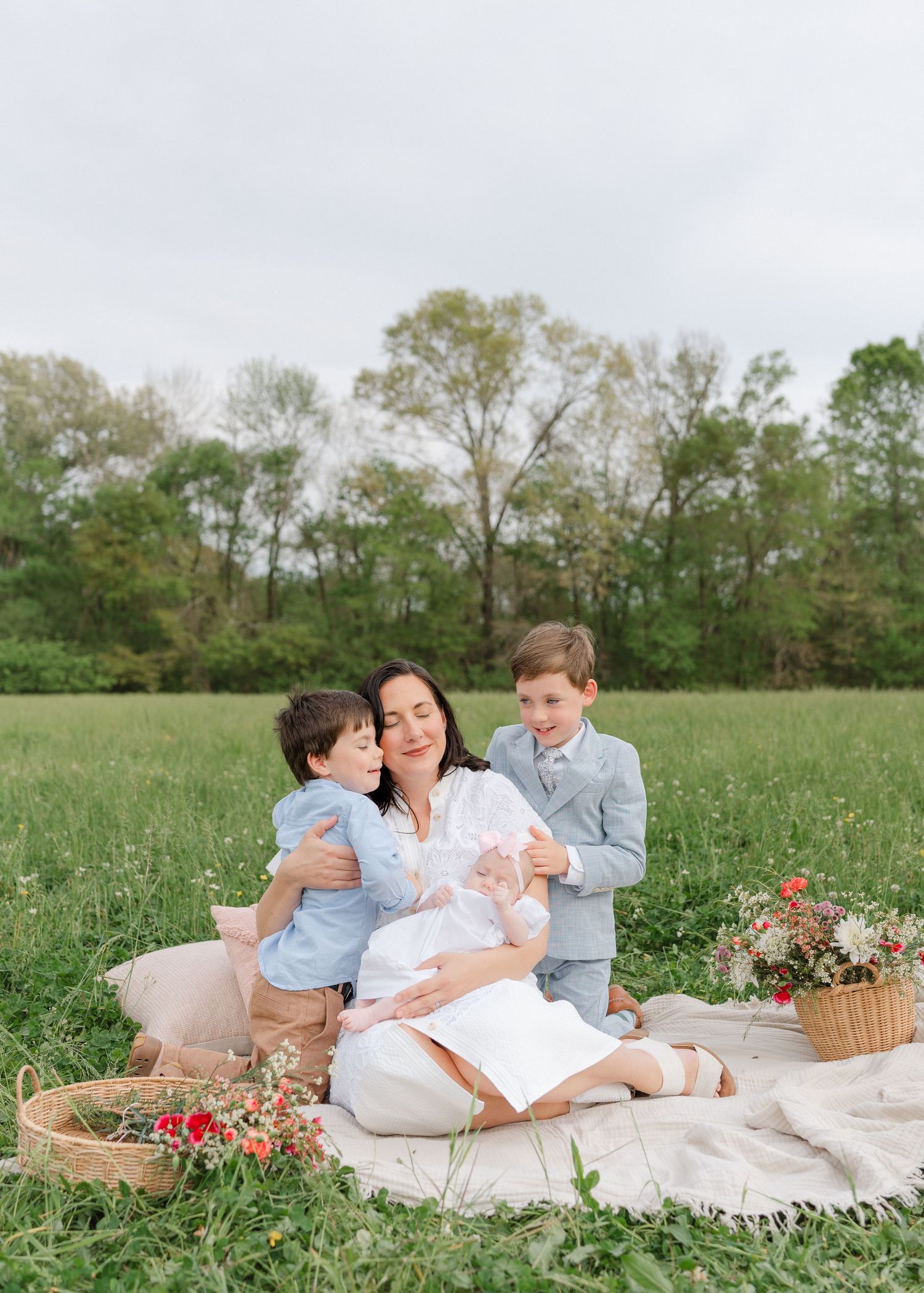 A mother sits on a blanket in a green field with her two young sons and newborn baby surrounded by flowers.