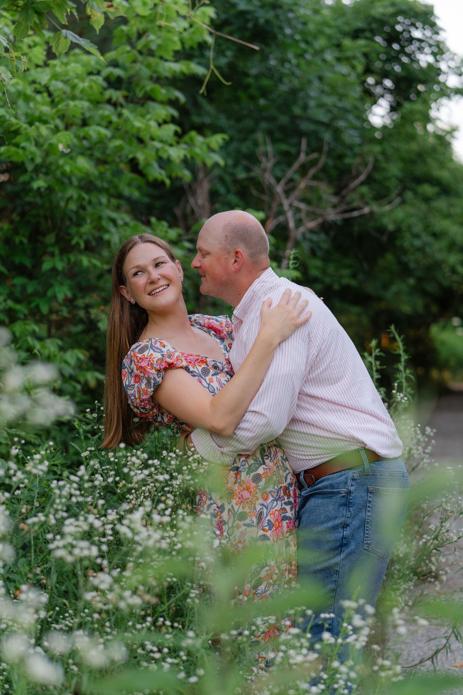 A smiling couple embraces among wildflowers and lush greenery, the woman in a floral dress laughing joyfully outdoors.