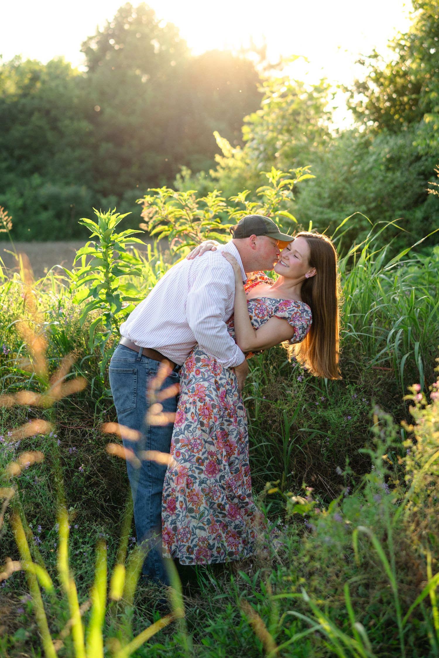 Couple sharing a romantic kiss in a sunlit garden, woman in floral dress embraced by man in white shirt and jeans.