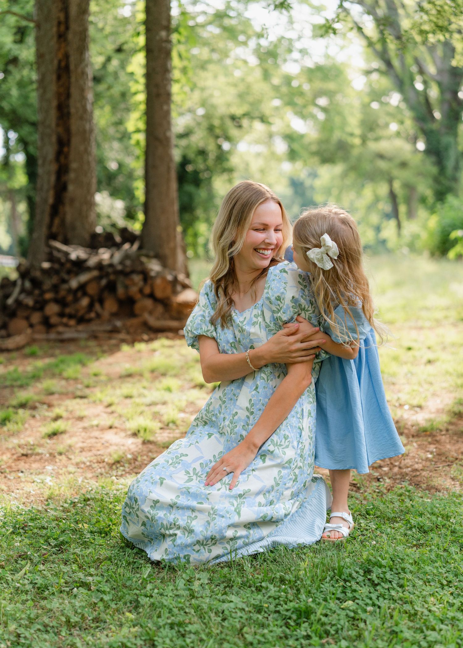 A smiling mother in a floral dress crouches to hug her young daughter in a blue dress outdoors.