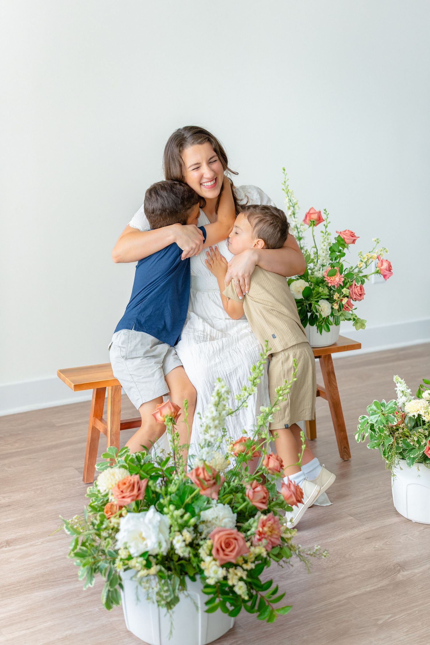 A laughing mother in a white dress is hugged by her two sons beside colorful flower bouquets.