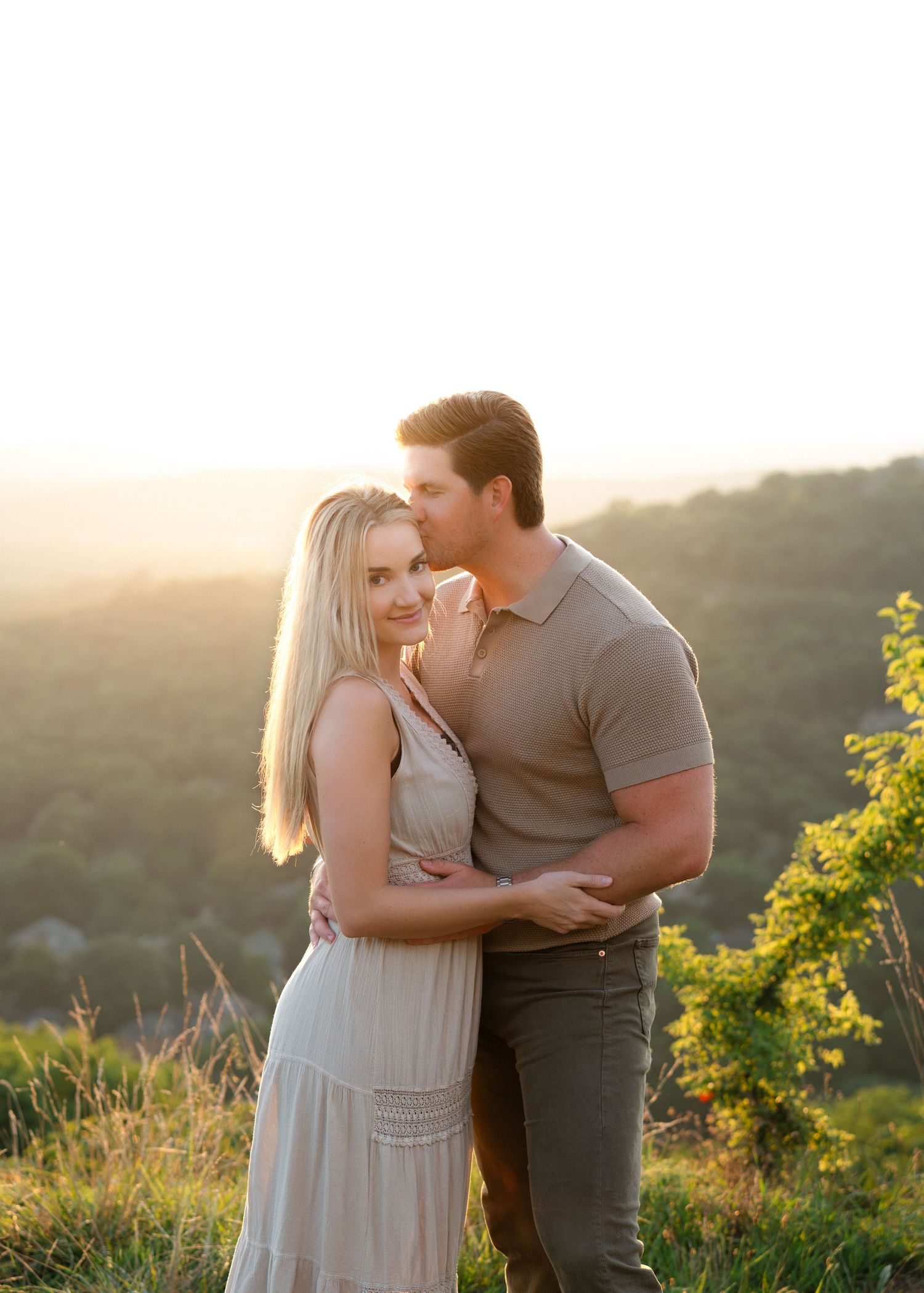Couple embracing at golden hour on a scenic overlook, woman in flowy dress smiling, man in olive shirt kissing her cheek.