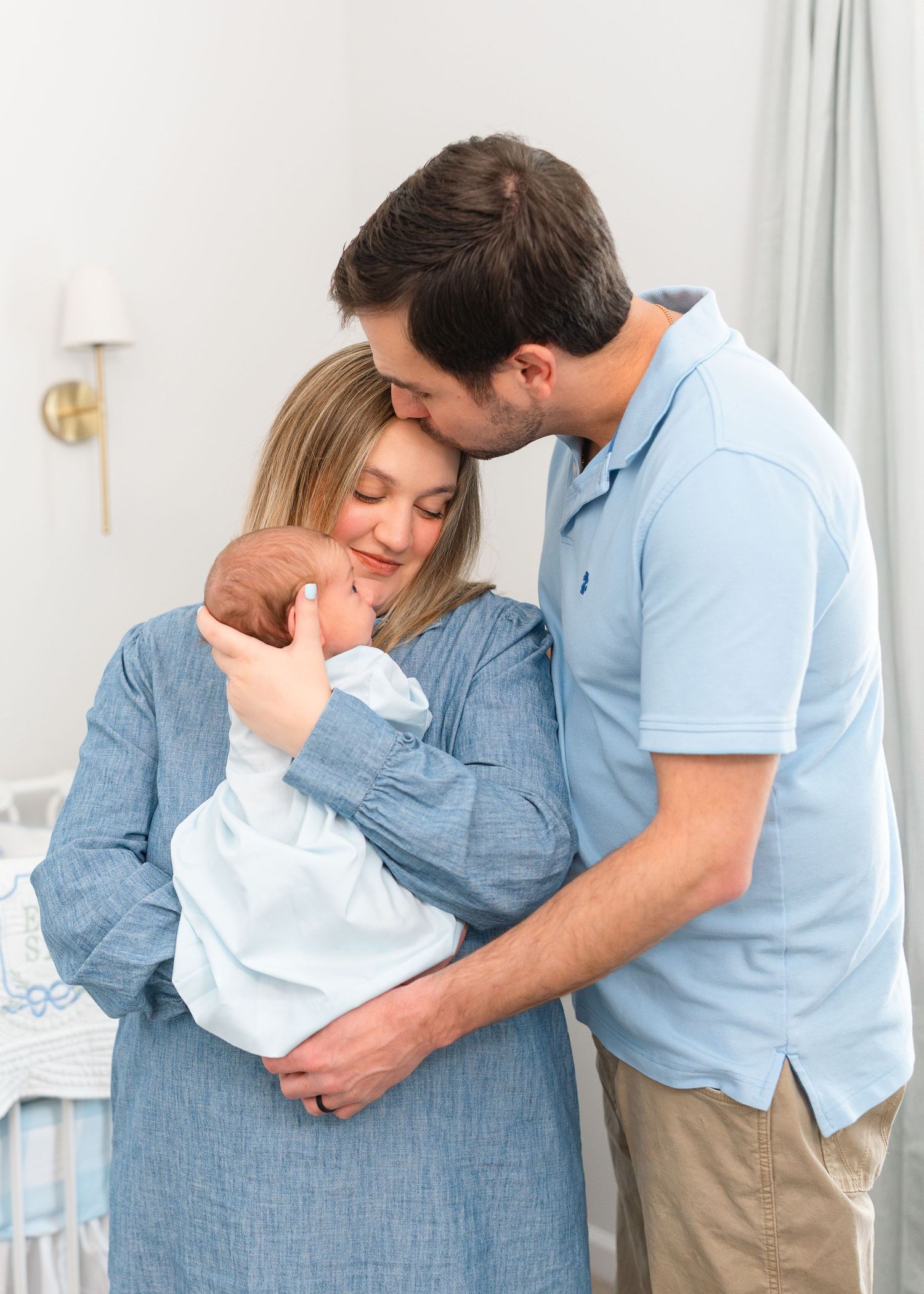 Parents in blue outfits tenderly holding newborn baby together in soft nursery light.