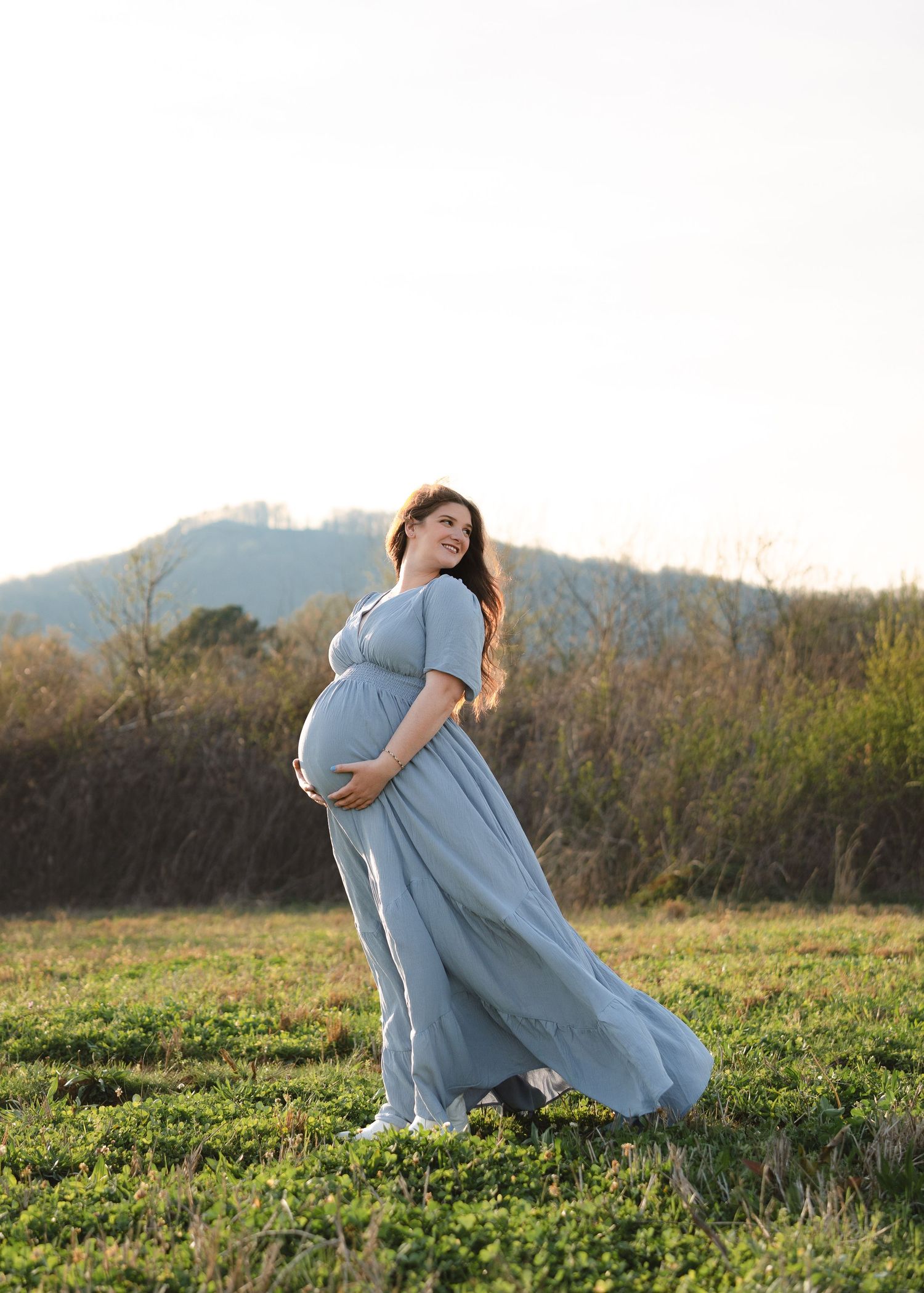 A pregnant woman in a flowing blue-gray gown stands in a sunlit green field with mountains behind her.