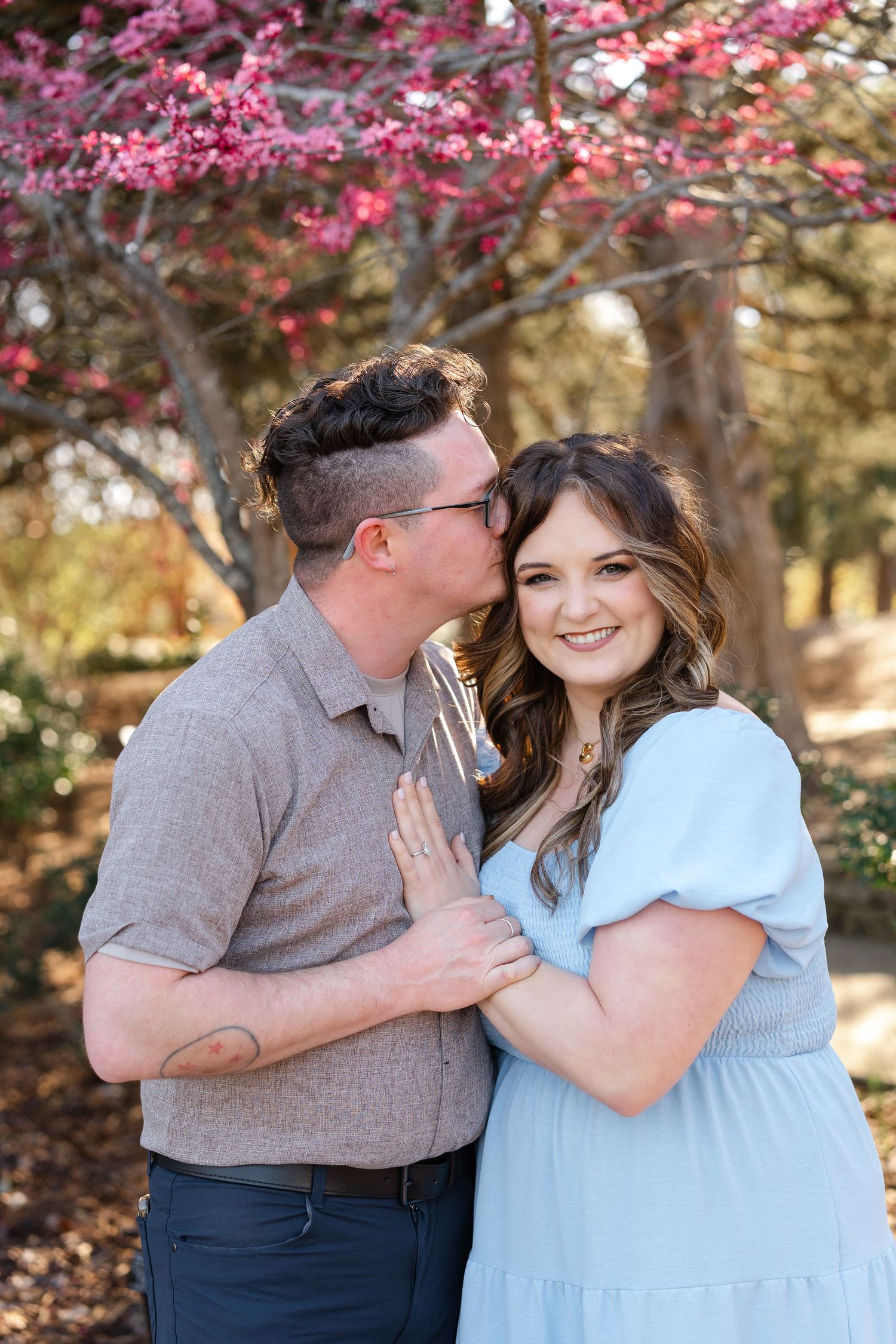 A smiling pregnant woman in a light blue dress is kissed on the cheek by her partner under a blooming tree.