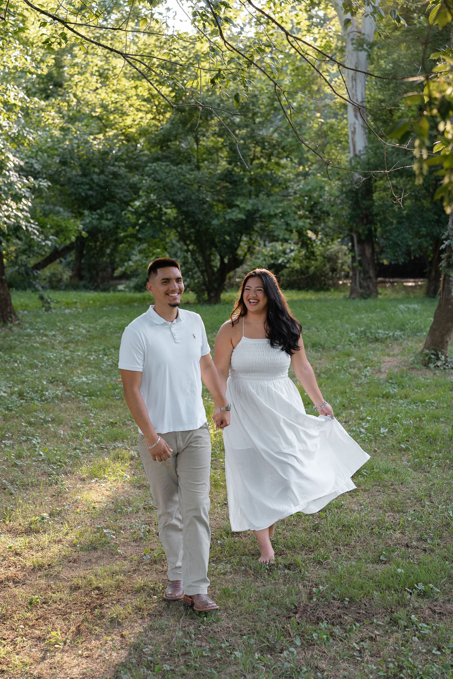 Couple in white outfits walking hand in hand through a lush green park, smiling joyfully on a sunny day.