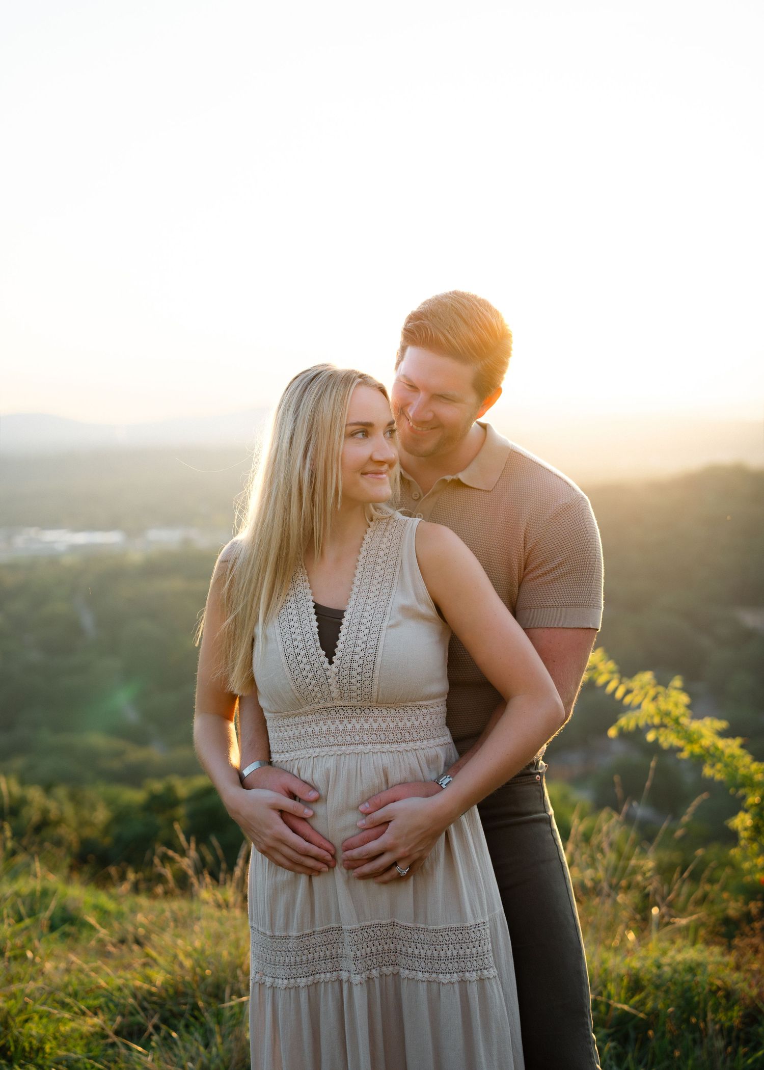 Couple embracing at golden hour sunset on a hilltop, pregnant woman in beige maxi dress with partner holding her belly.