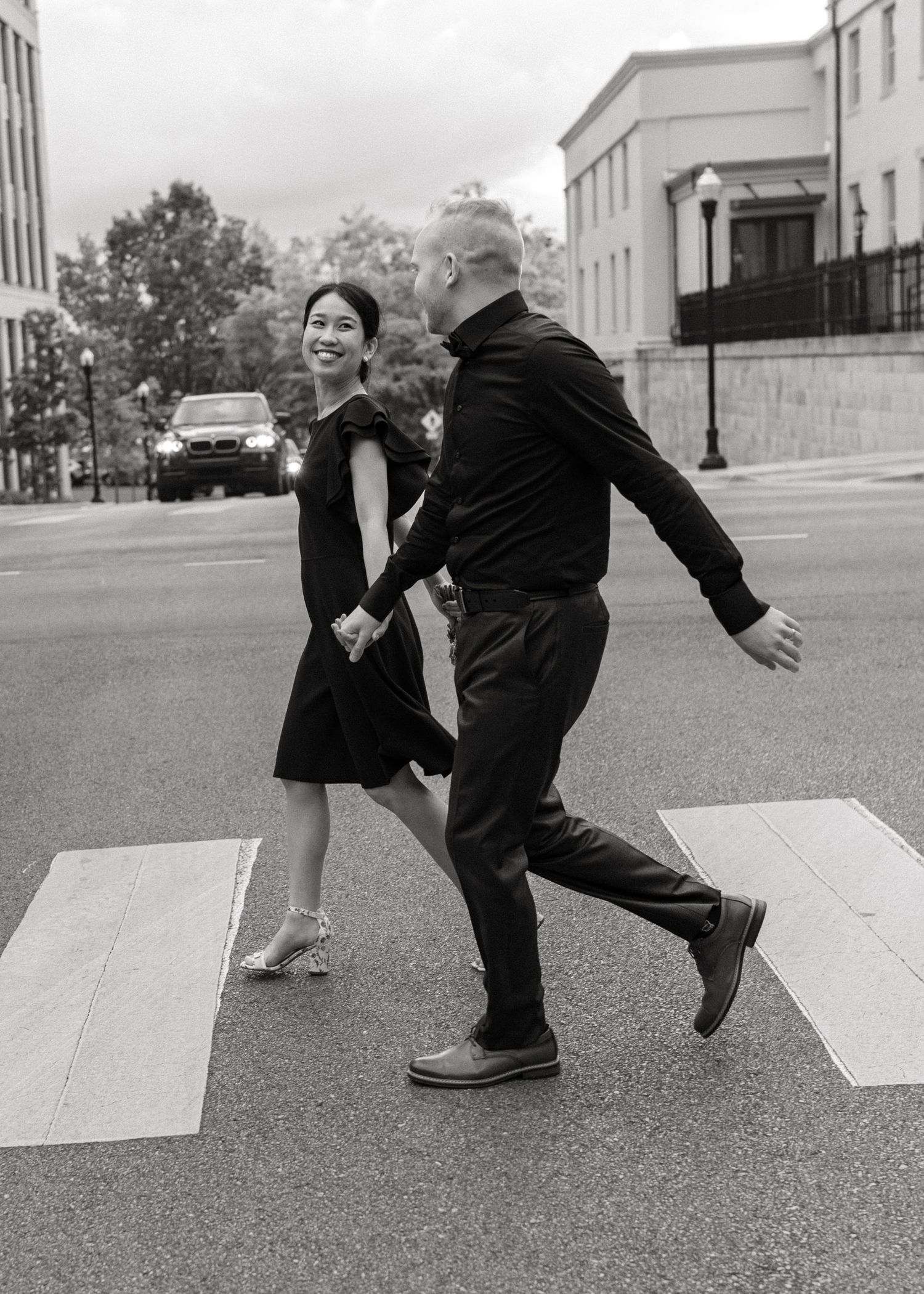 Couple in black outfits holding hands and smiling while crossing a zebra crossing on a city street in black and white.