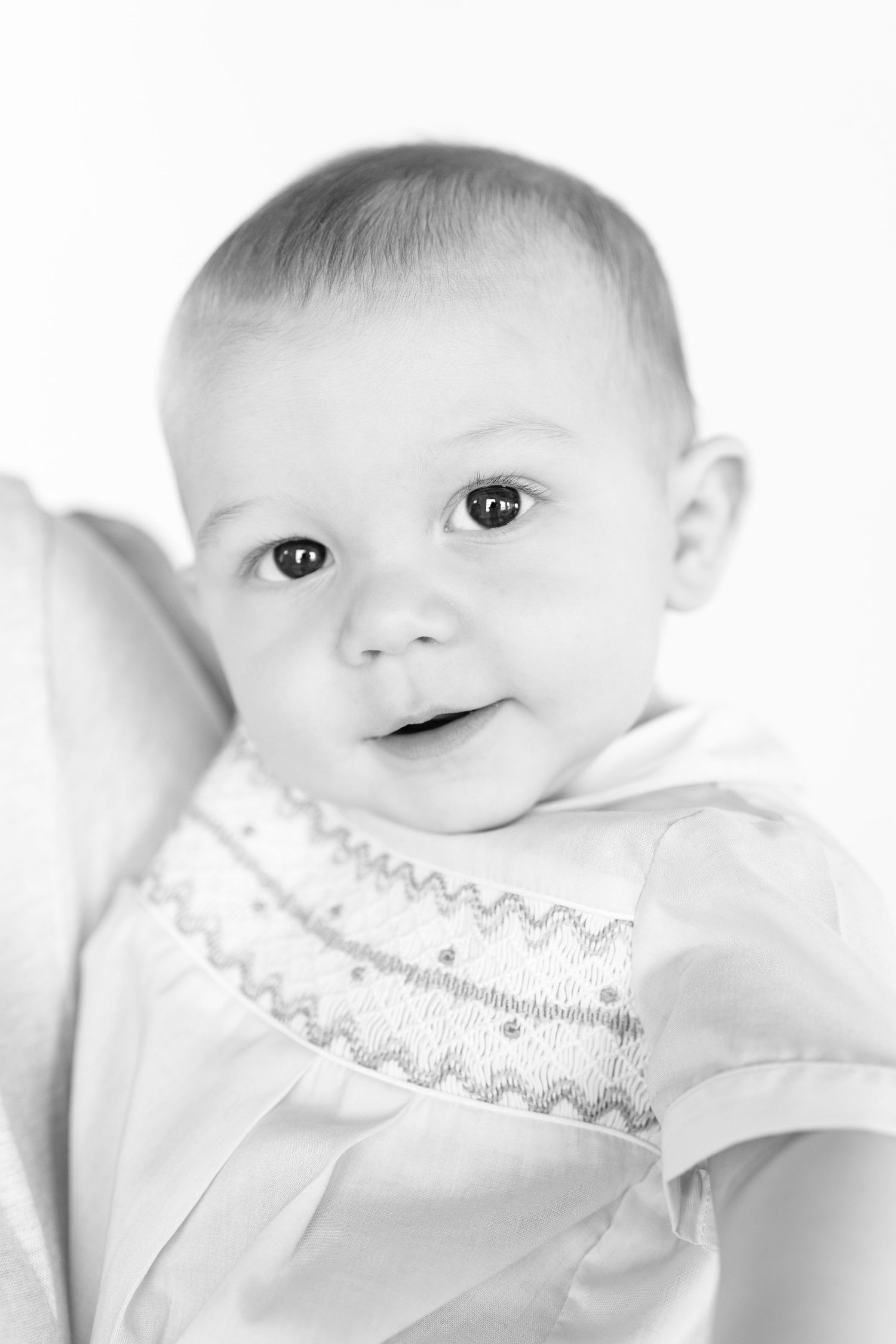 Black and white portrait of an adorable baby in a smocked white outfit, looking at the camera with bright eyes.