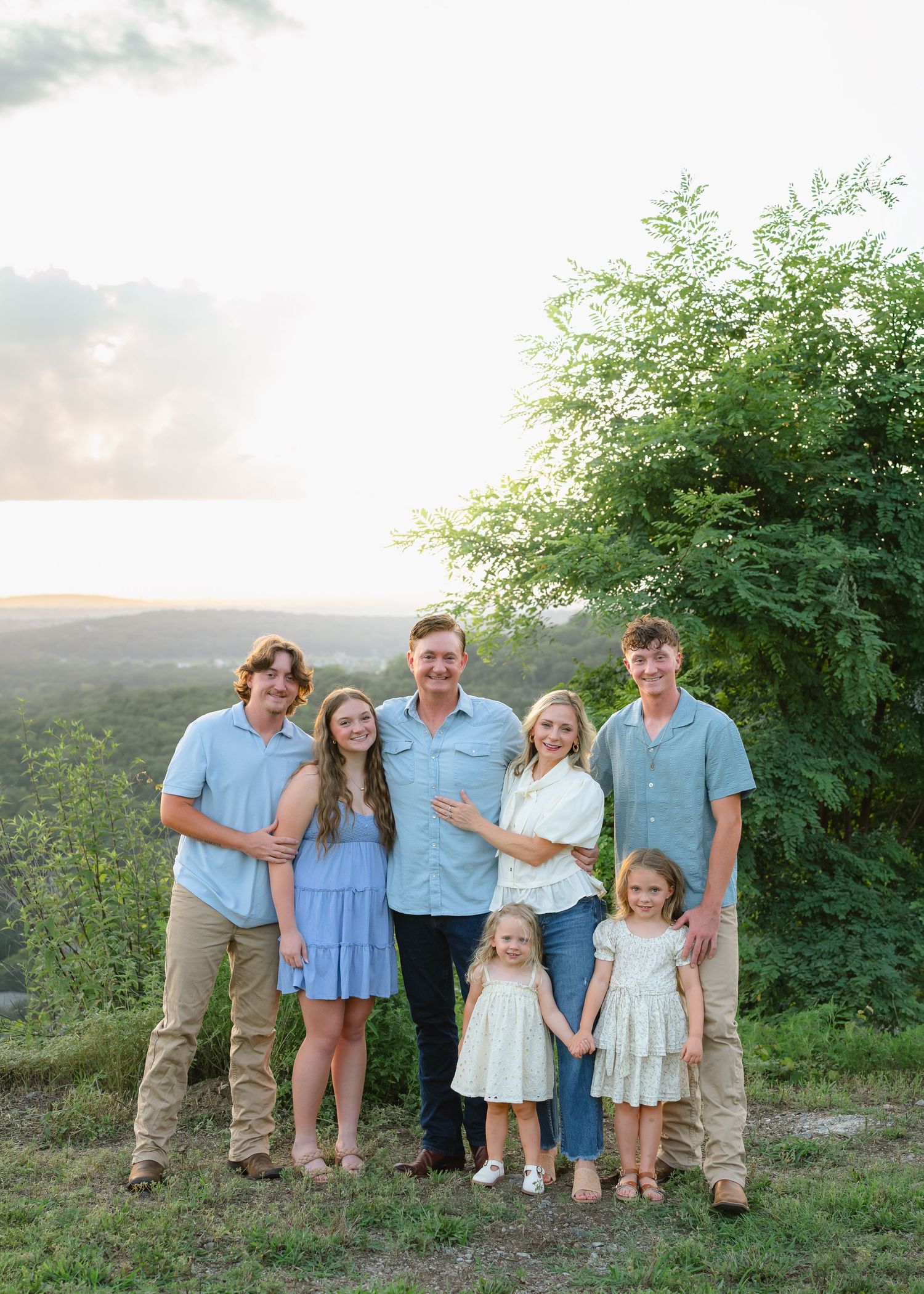 Family of seven posing outdoors at golden hour with lush green trees and scenic landscape in the background.