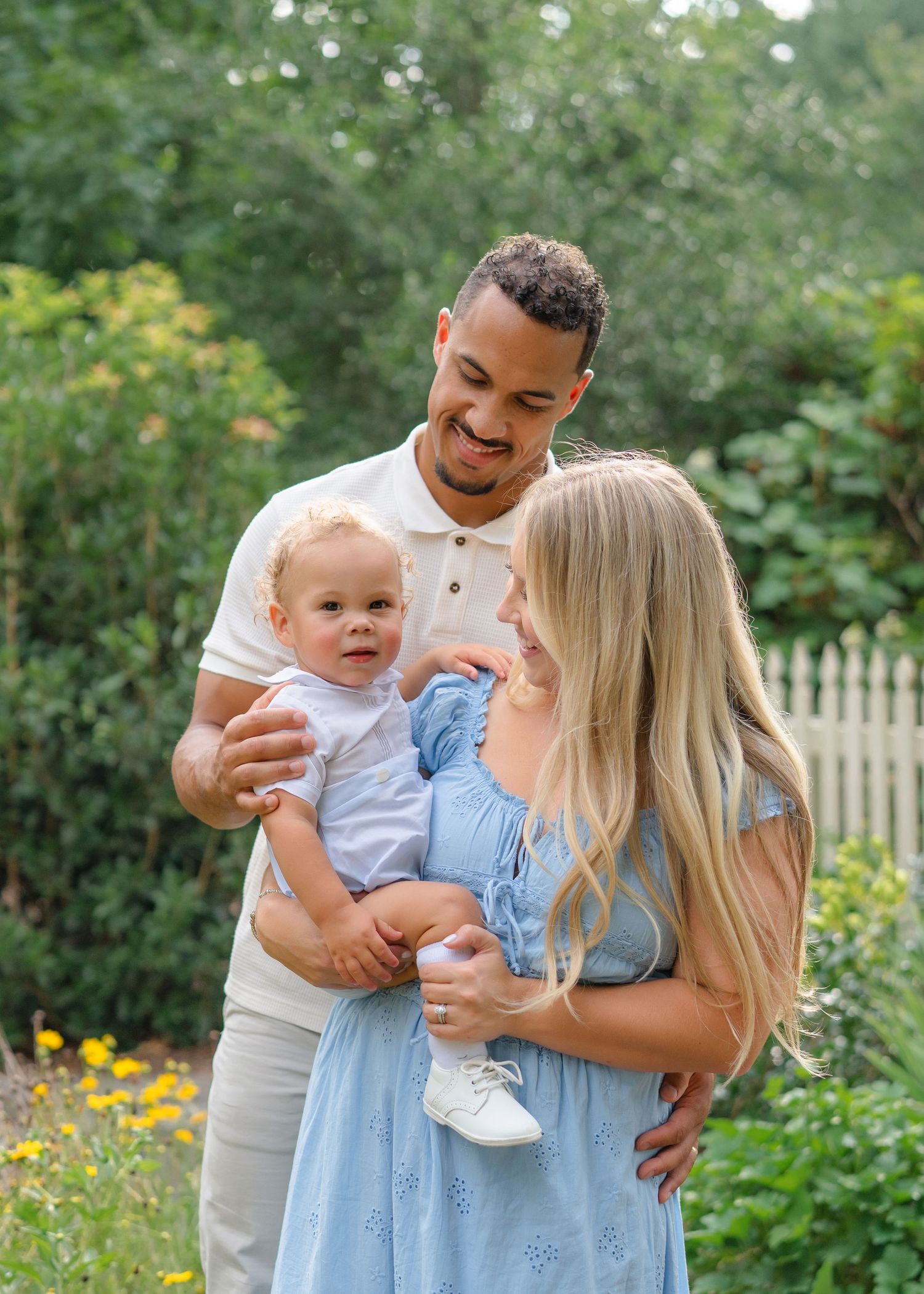 A happy young family with a baby boy poses outdoors in a lush garden setting on a sunny day.