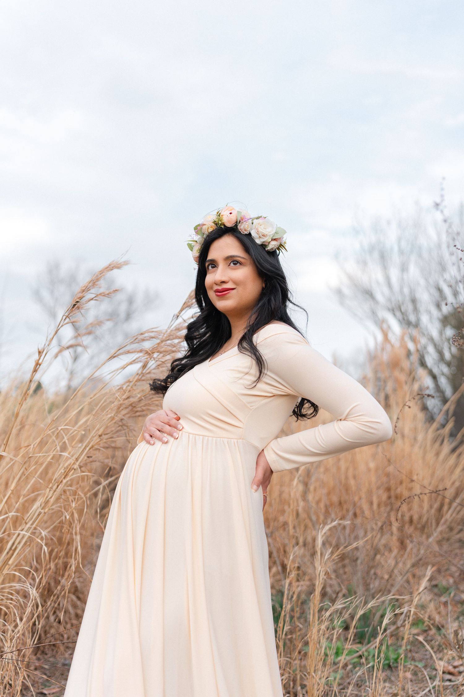 Pregnant woman in cream maternity dress and floral crown poses in tall grass field.