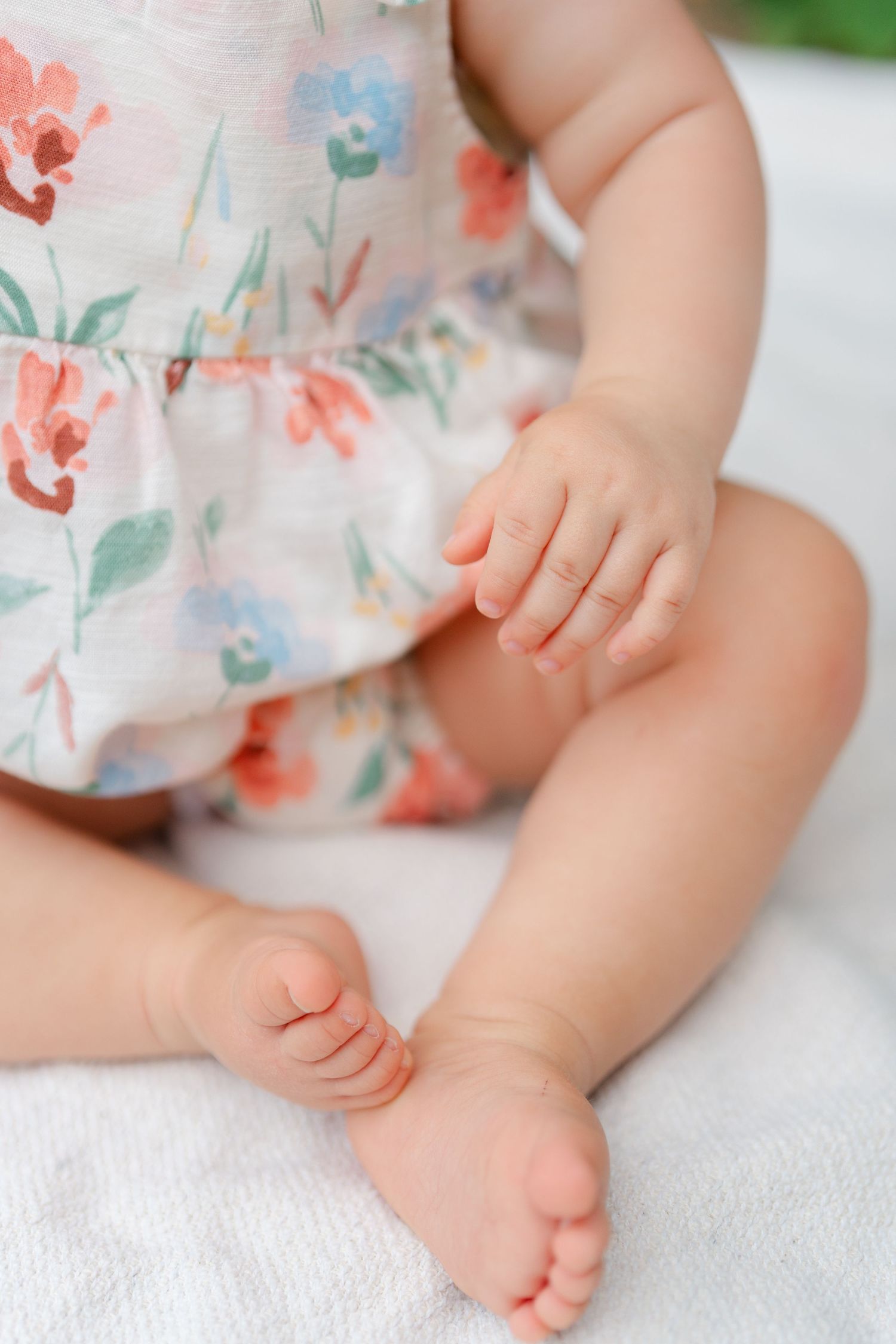 Close-up of a baby's tiny hands and feet wearing a floral romper, sitting on a white blanket.