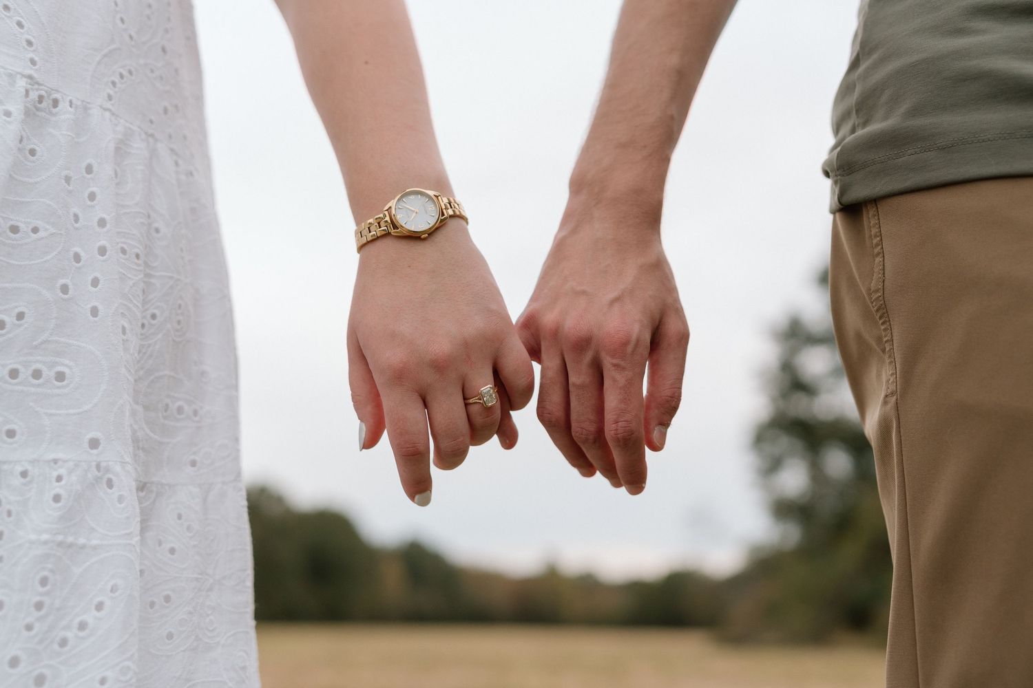 Couple gently holding fingertips together outdoors, woman in white dress with gold watch and engagement ring.