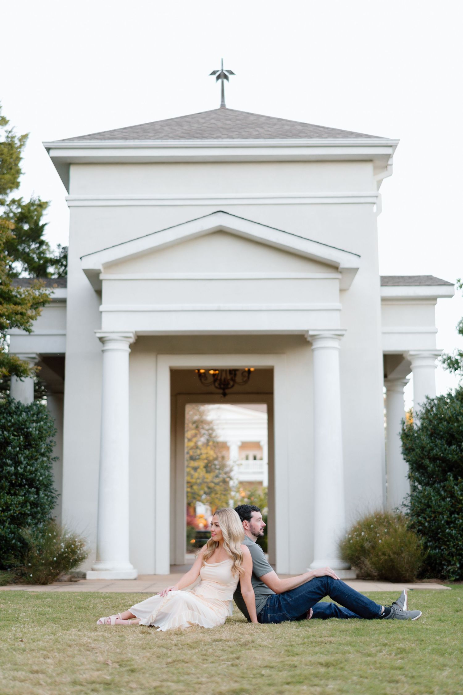 Engaged couple sitting back-to-back on lawn in front of elegant white columned building at dusk.