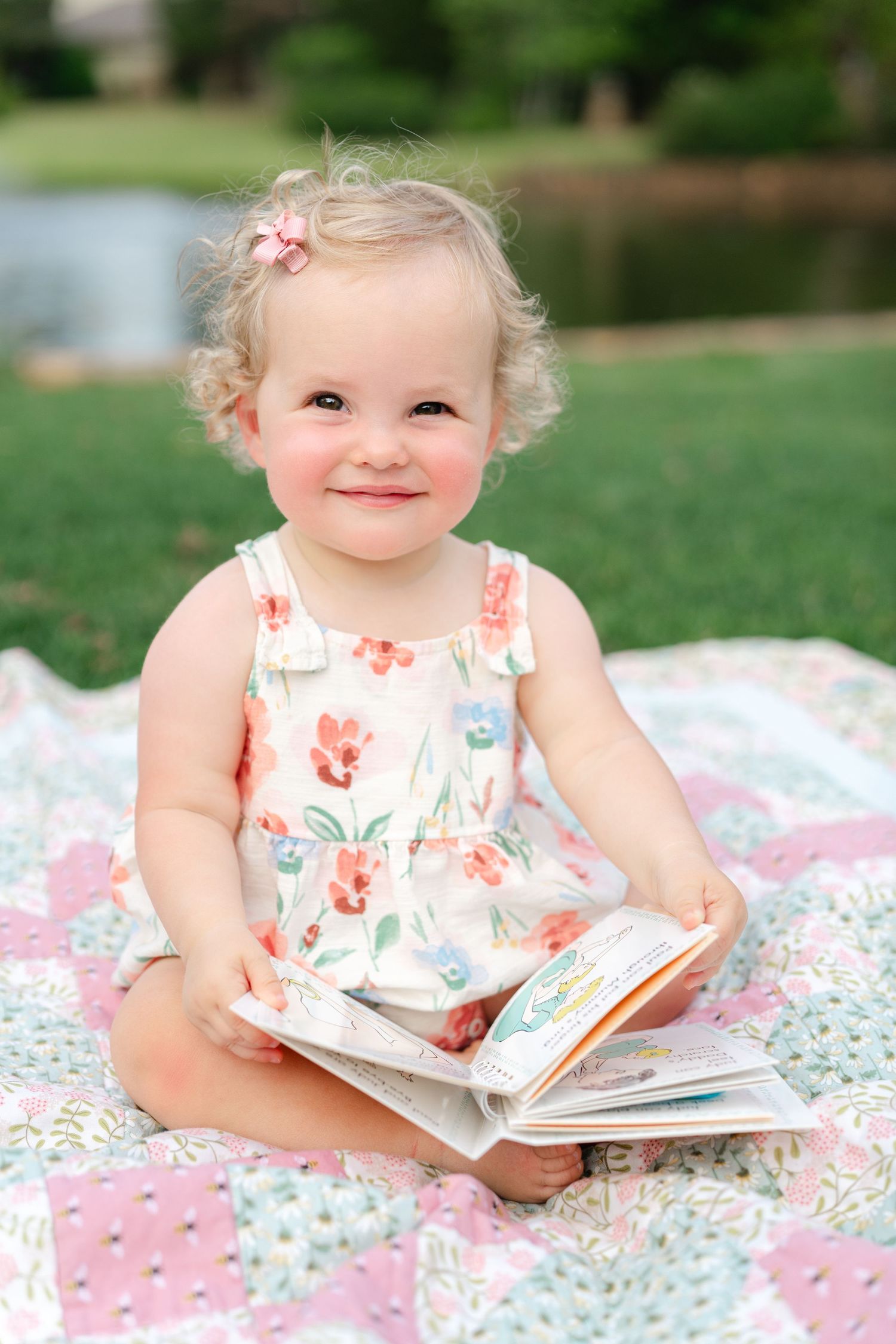 Smiling toddler girl in floral dress sitting on quilt outdoors holding a book.