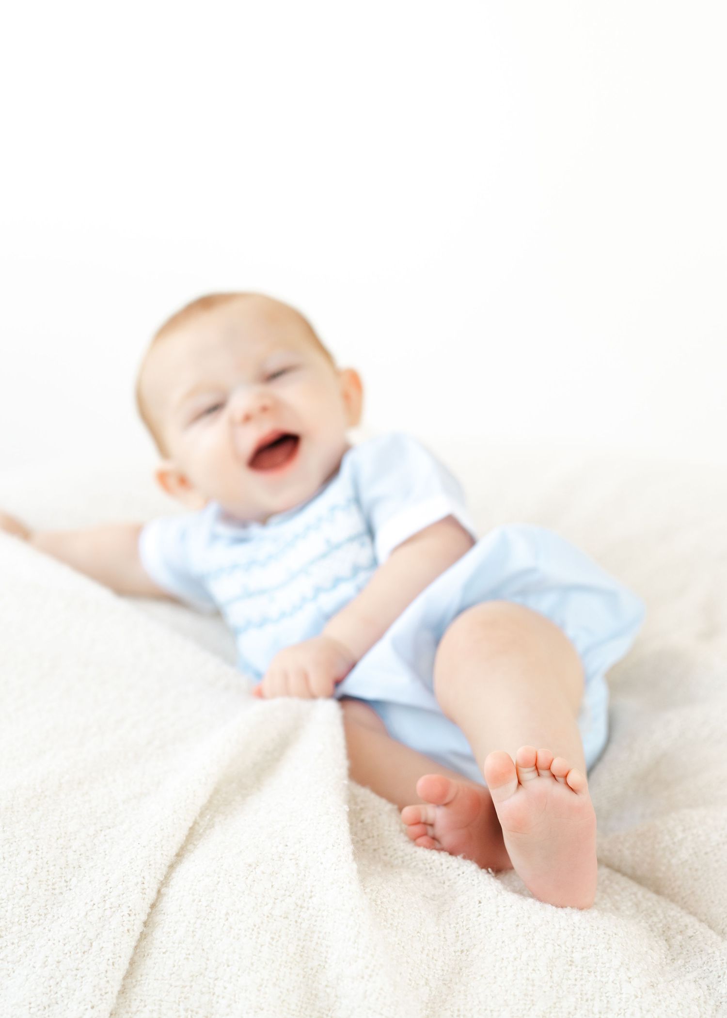 Happy baby in blue onesie laughing while sitting on a white blanket, with tiny bare feet in the foreground.