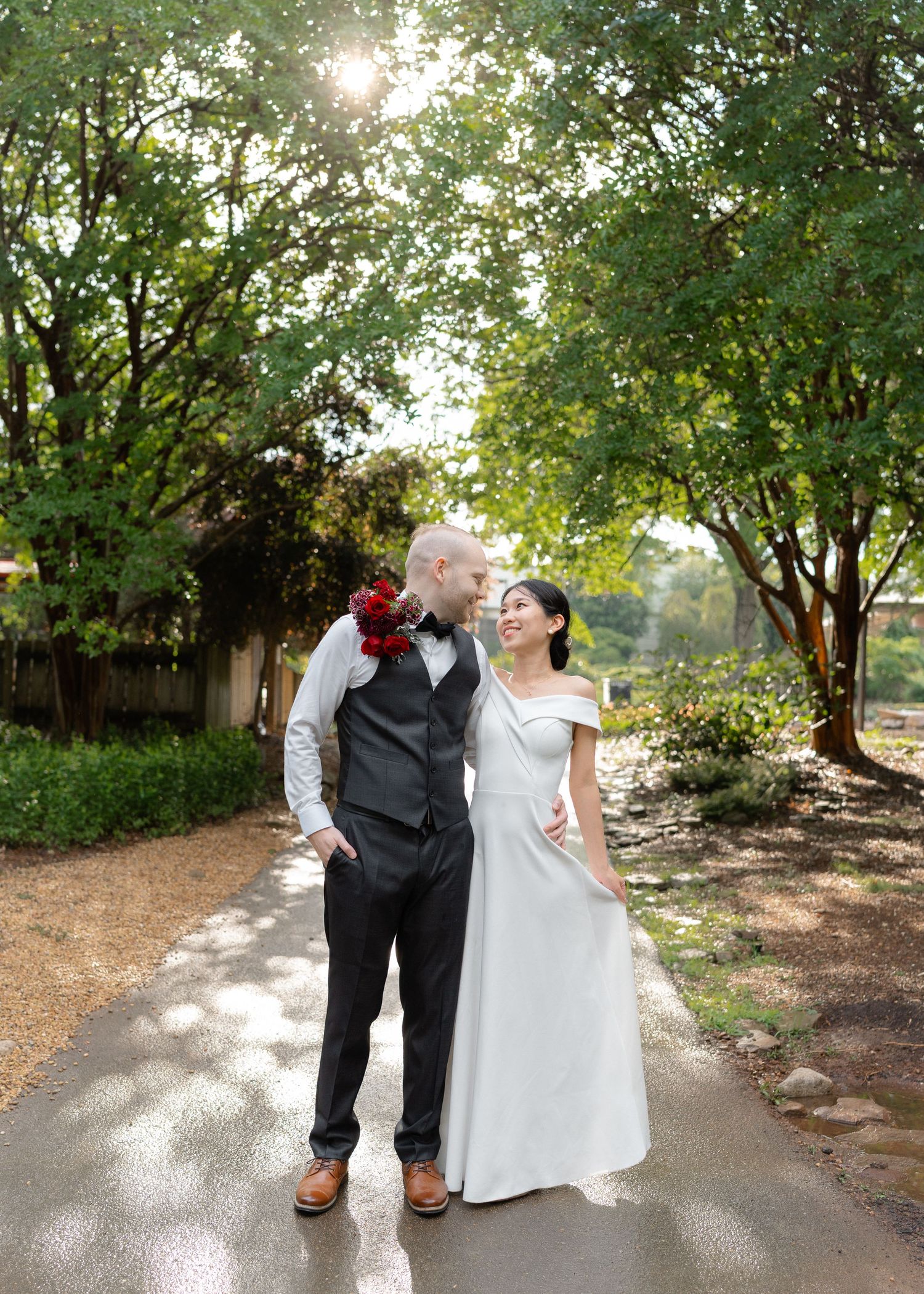 Bride in white gown and groom in gray vest share a romantic moment on a tree-lined garden path.