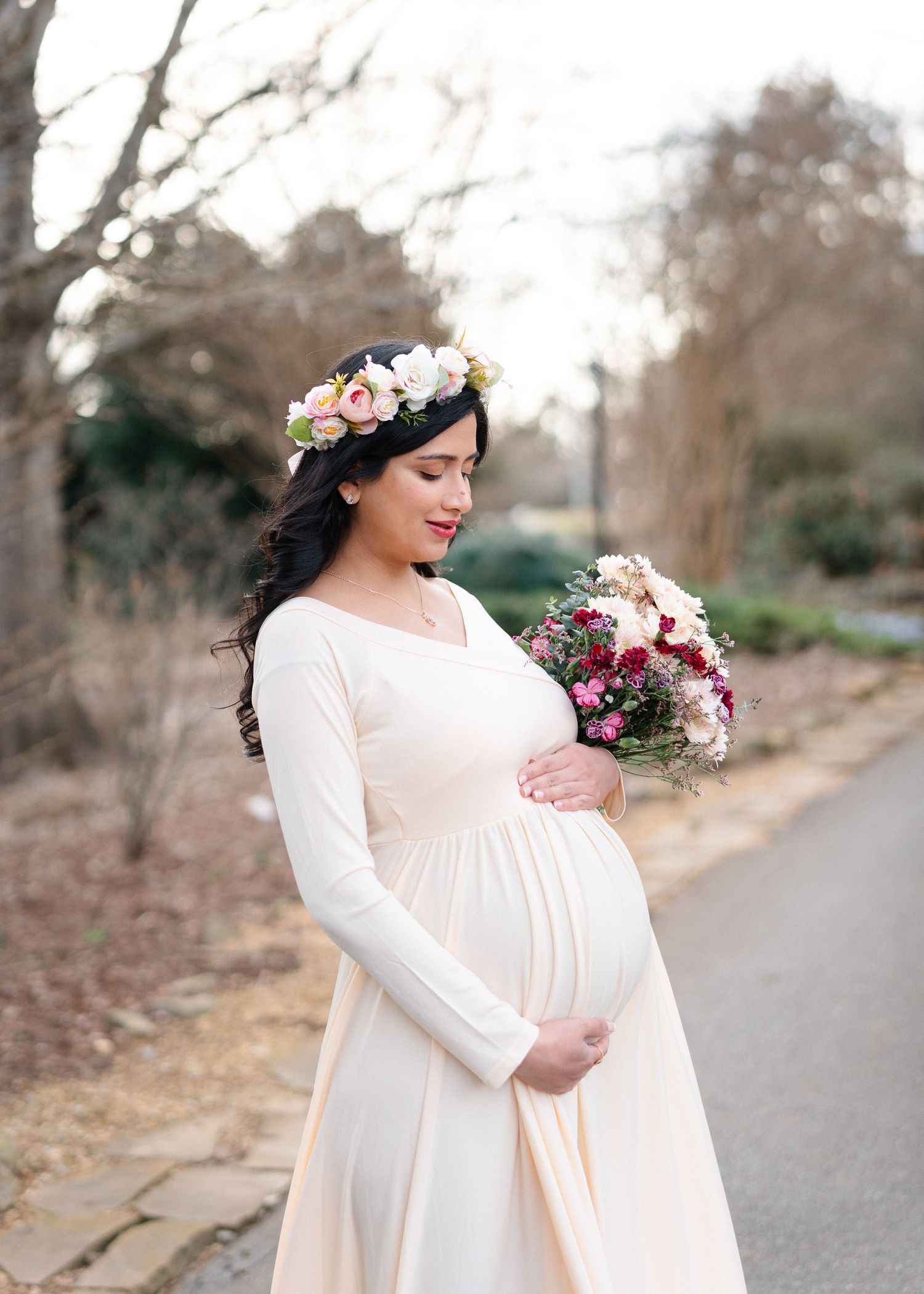 Pregnant woman in cream gown and floral crown holds bouquet outdoors.