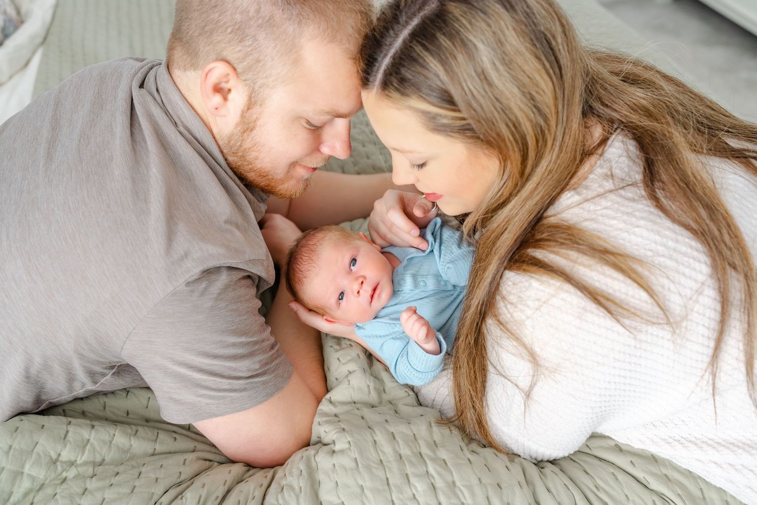 New parents lean in close together smiling at alert newborn baby in blue outfit on bed.