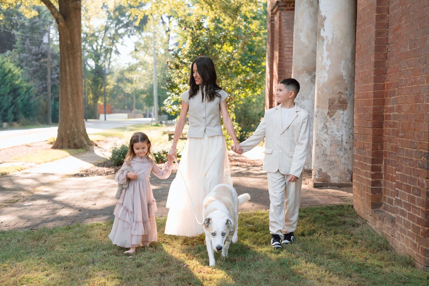 Mother walks with son, daughter, and white dog along sunny path beside brick building.