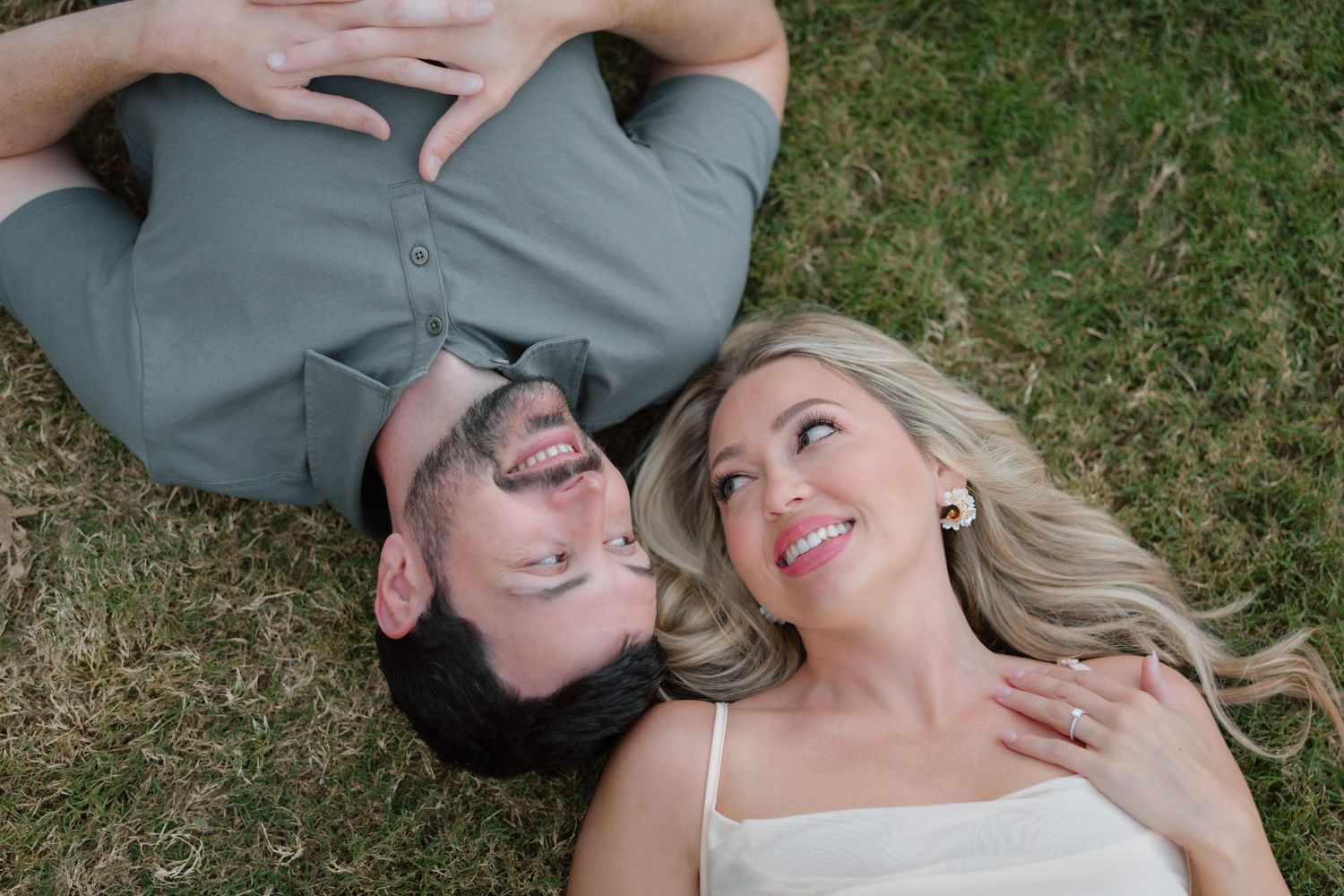 Happy couple lying on green grass looking up and laughing together during outdoor engagement session.