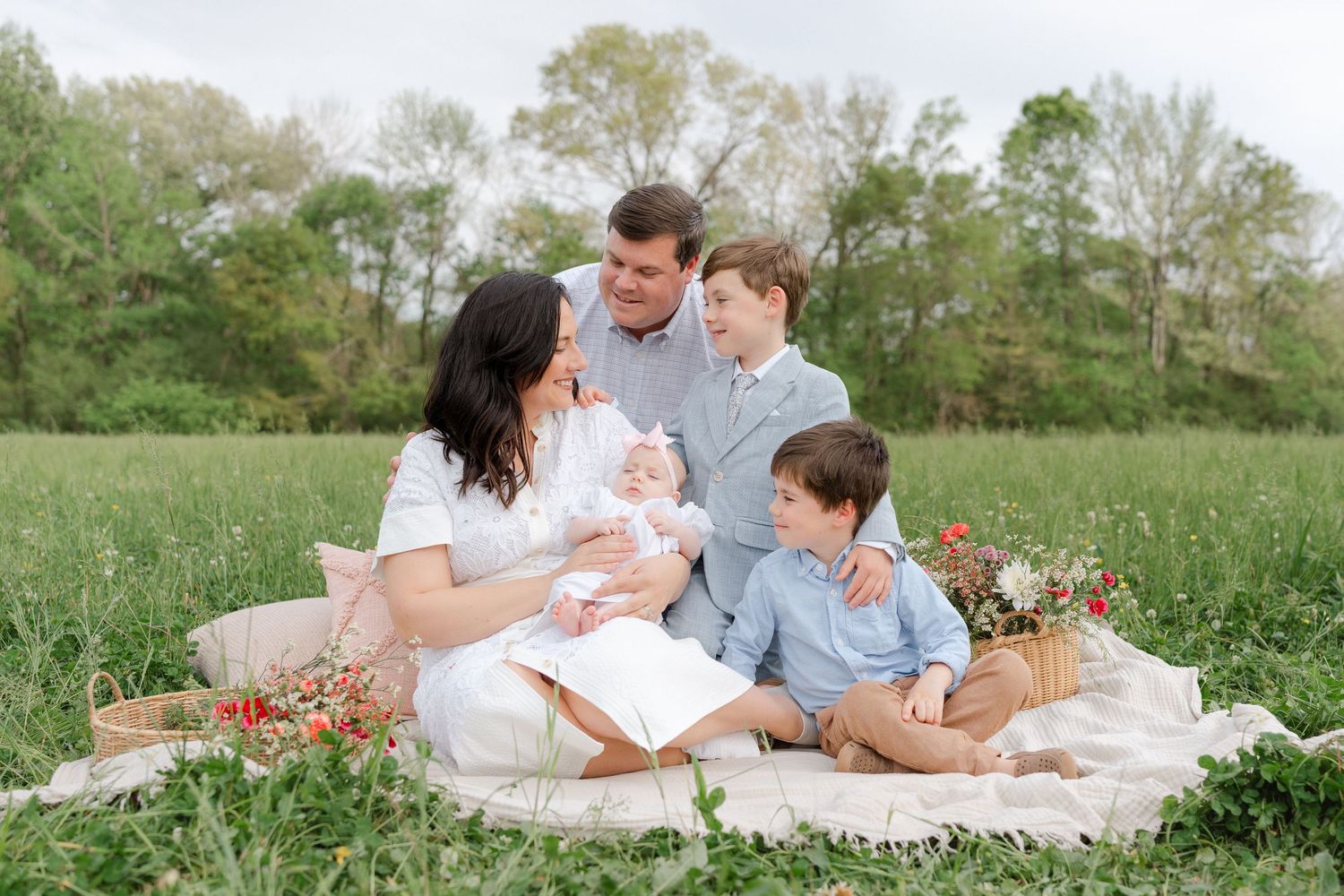Happy family of five sits together on a picnic blanket in a green field with flowers.