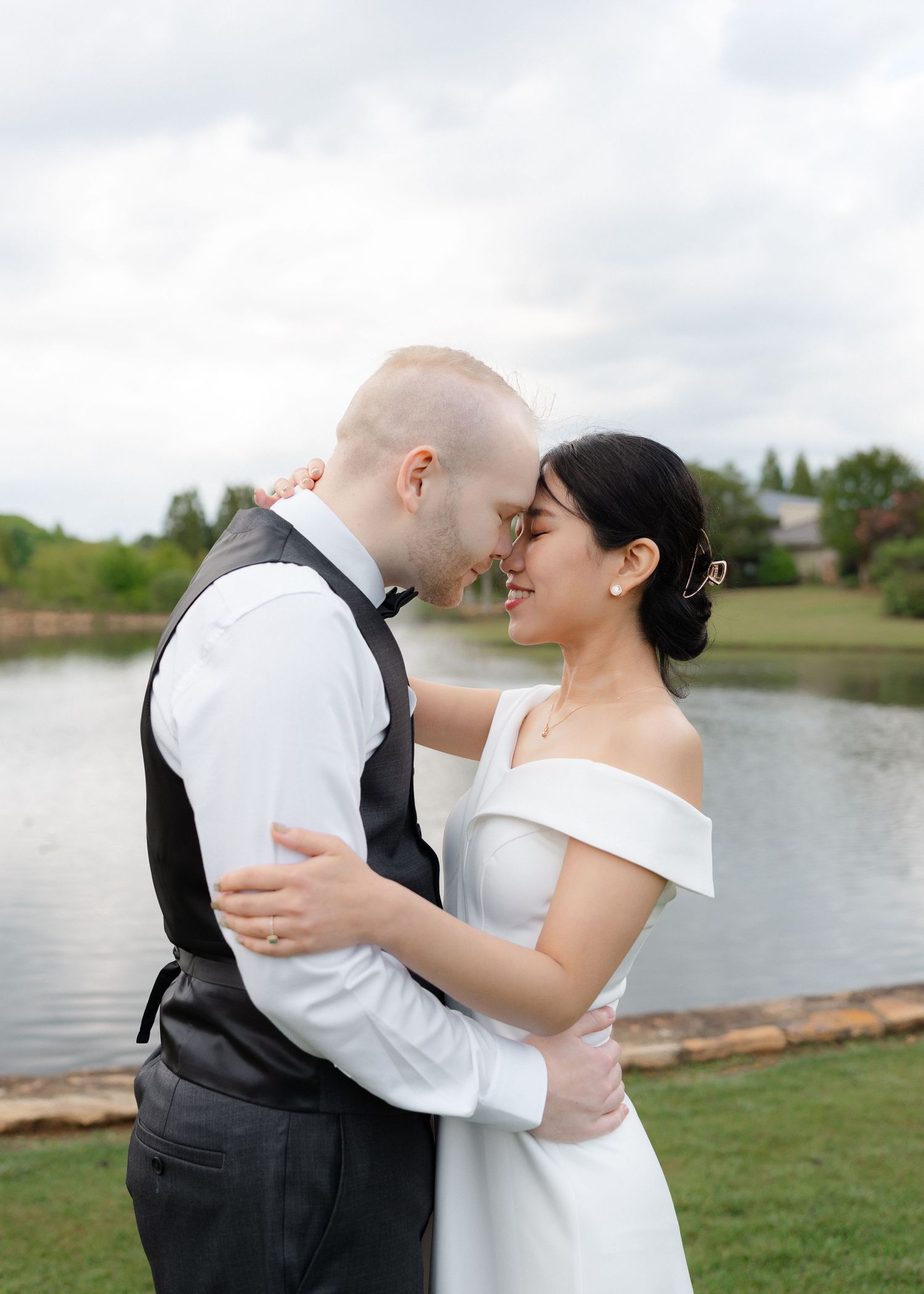 A couple shares a romantic moment in wedding attire by a serene lake, the bride in an off-shoulder gown.