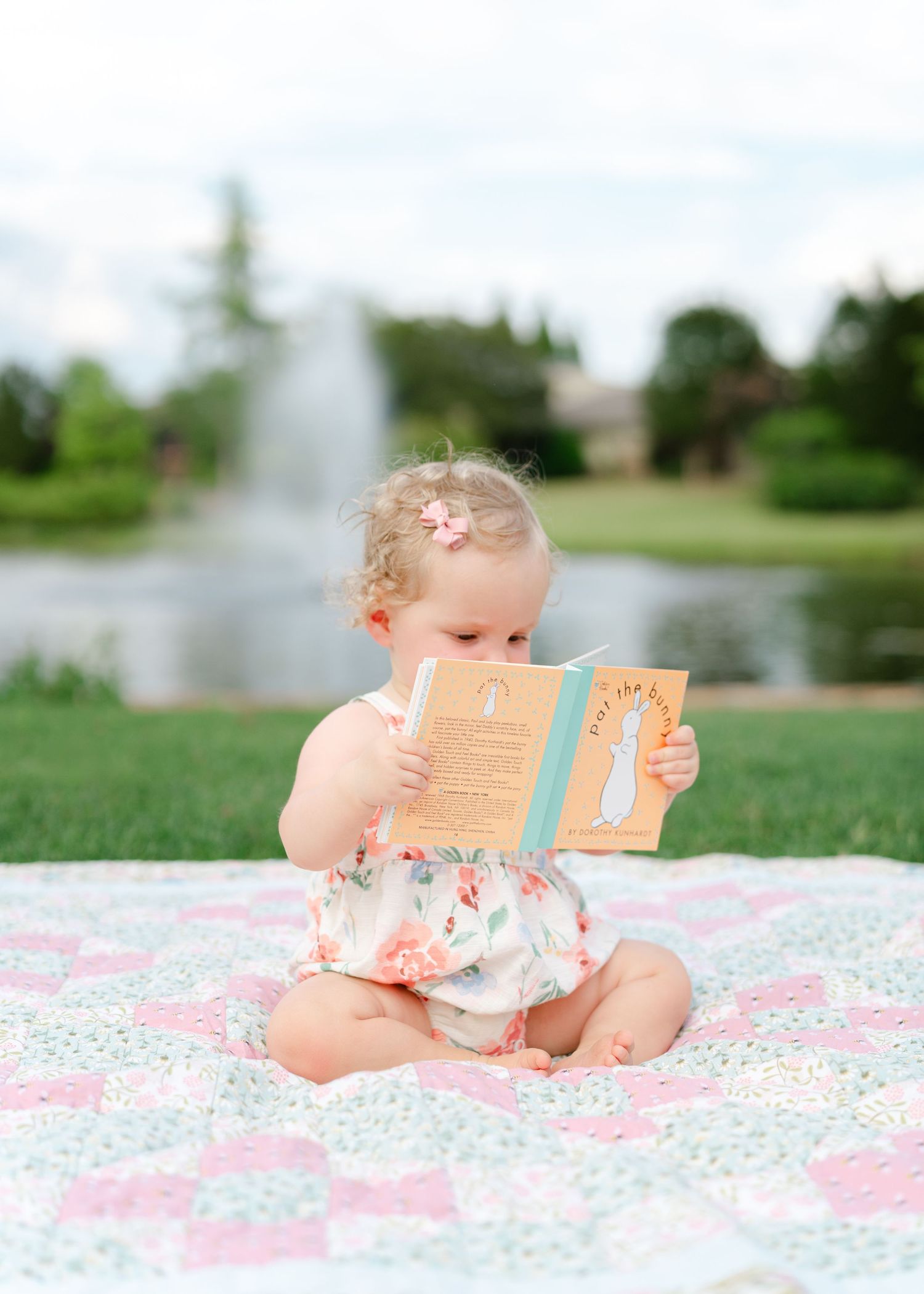 Adorable baby girl in floral outfit reading a book on a quilt blanket near a fountain in a park.