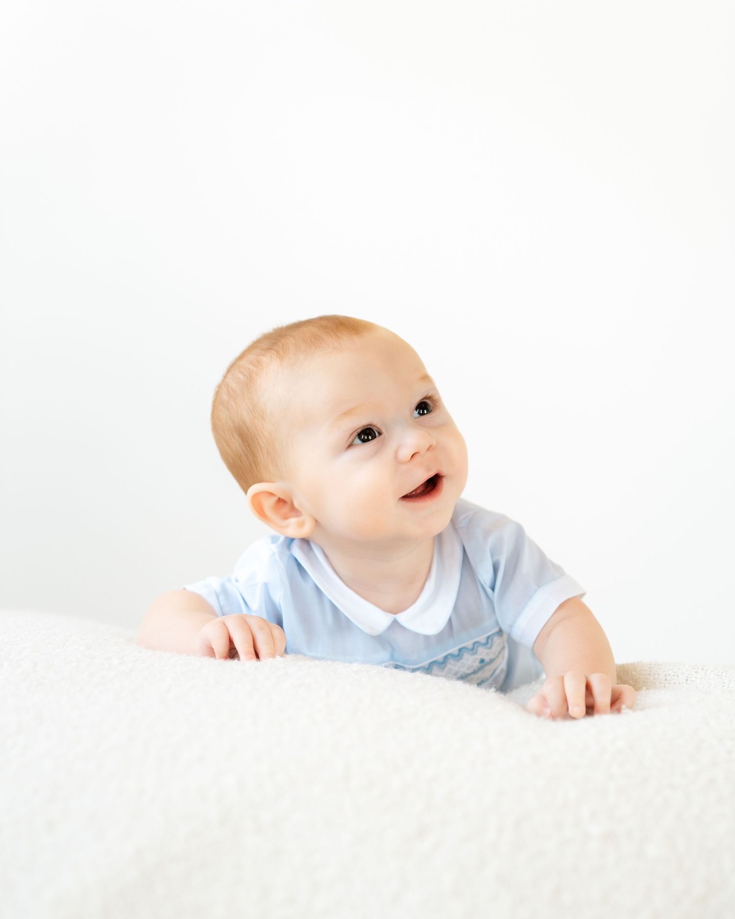 Smiling baby in a blue outfit doing tummy time on a white fluffy surface against a bright white background.