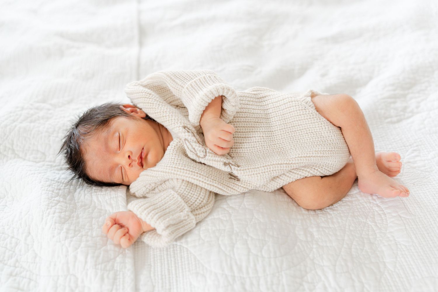 Peaceful newborn baby sleeps curled up in beige knit romper on white textured blanket.
