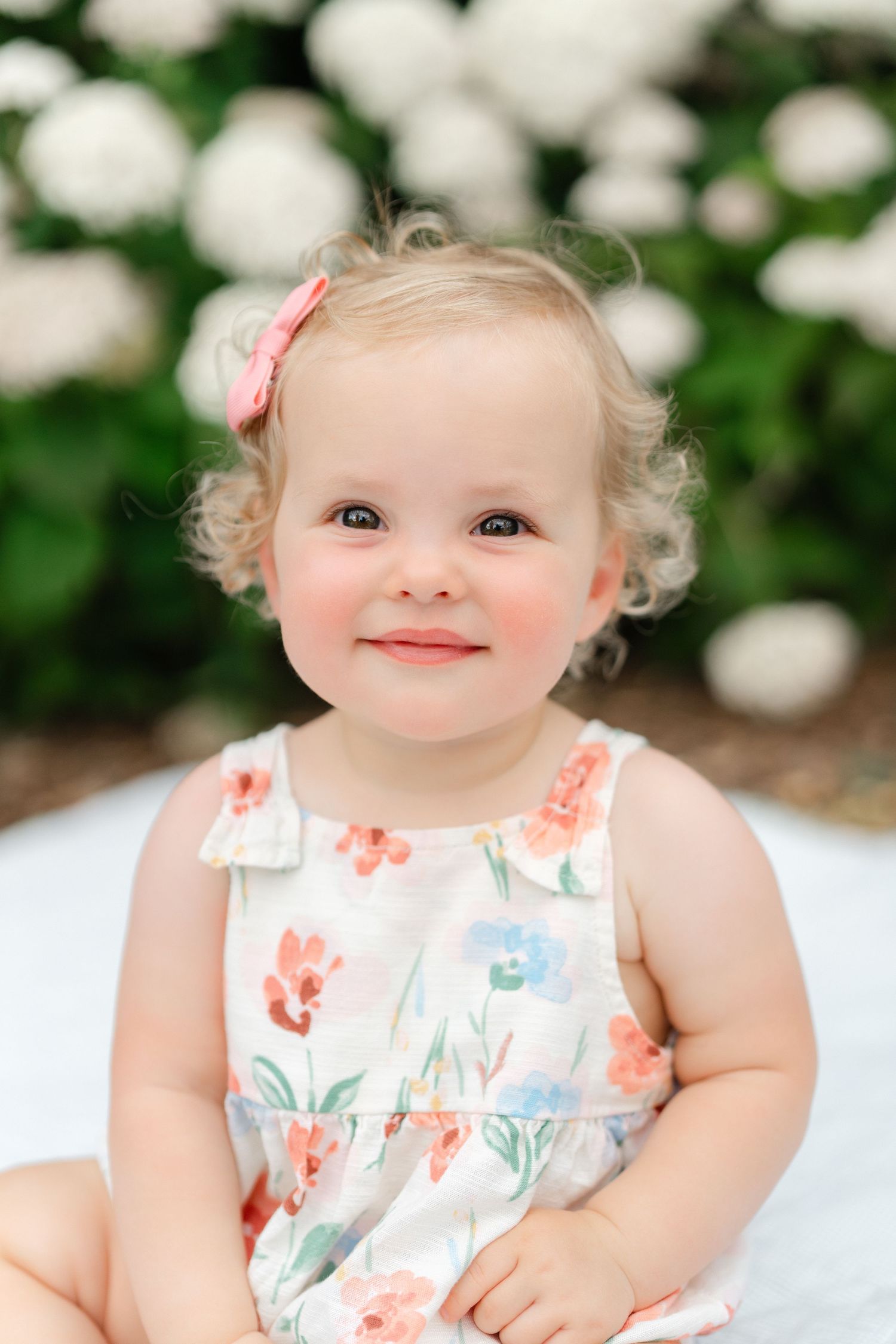 Adorable toddler girl with curly blonde hair in a floral dress smiling outdoors near white flowers.