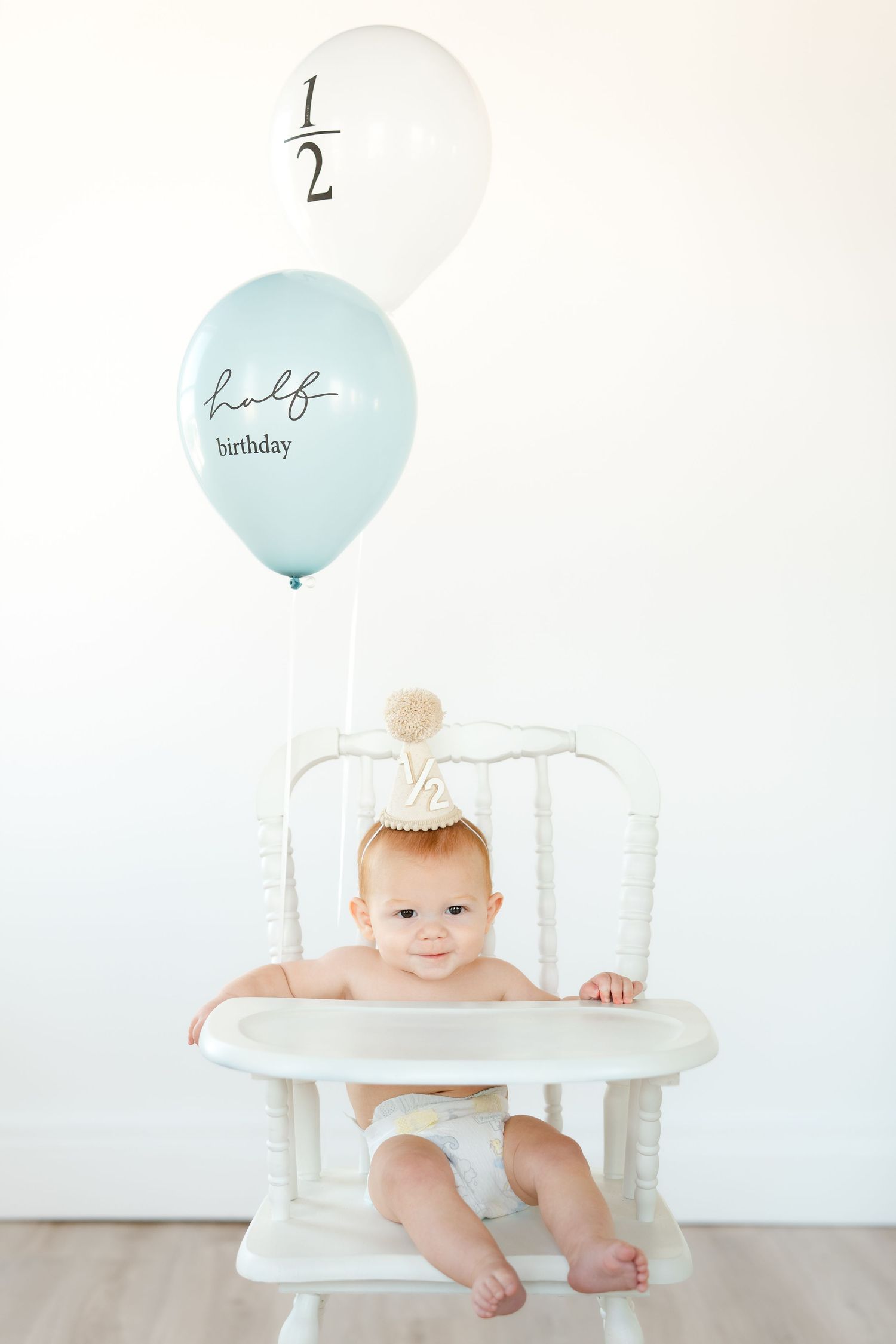 Smiling baby in white high chair wearing party hat with half birthday balloons for a 6-month celebration photo.