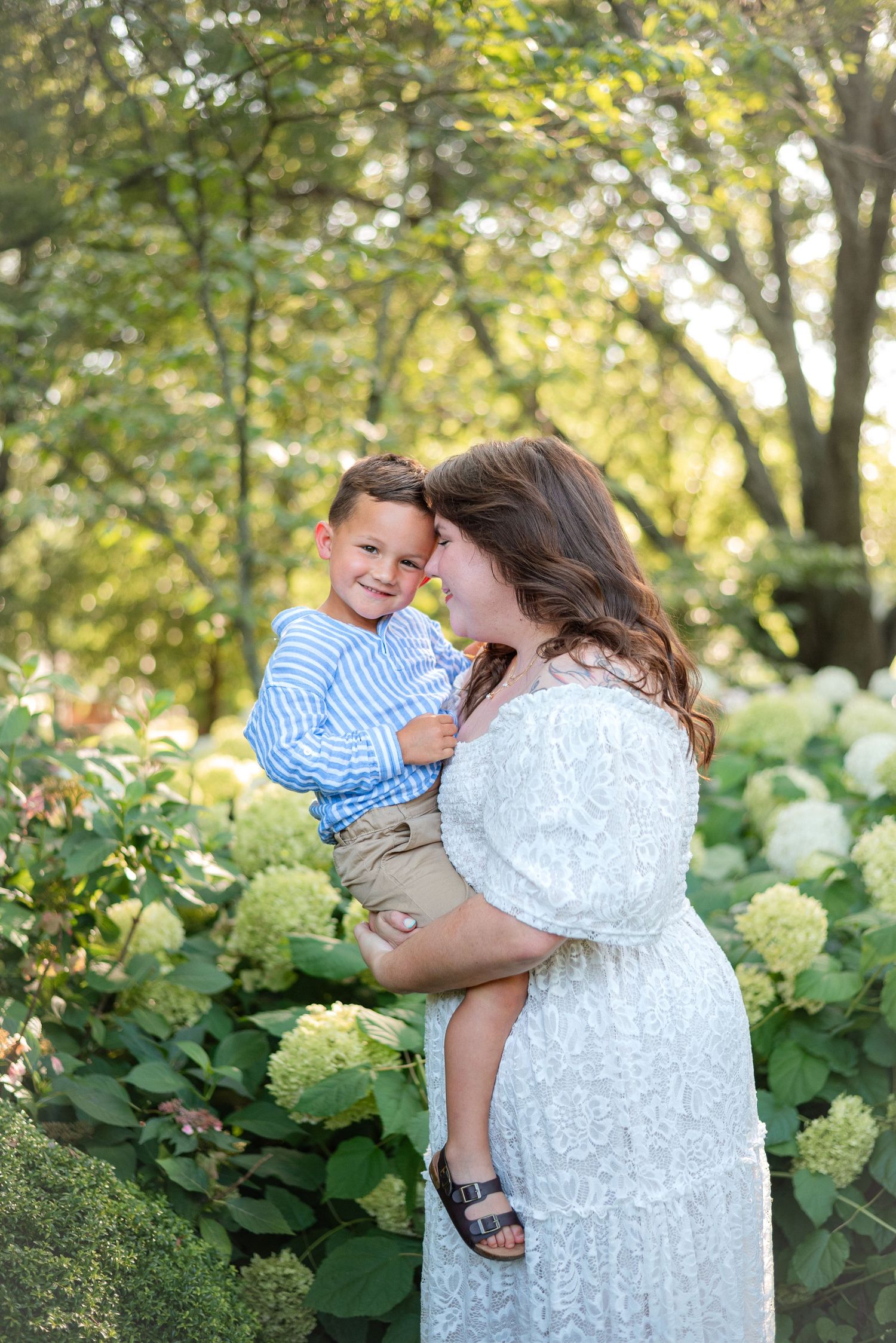 Smiling boy in blue stripes shares a joyful moment with mother among white hydrangeas.