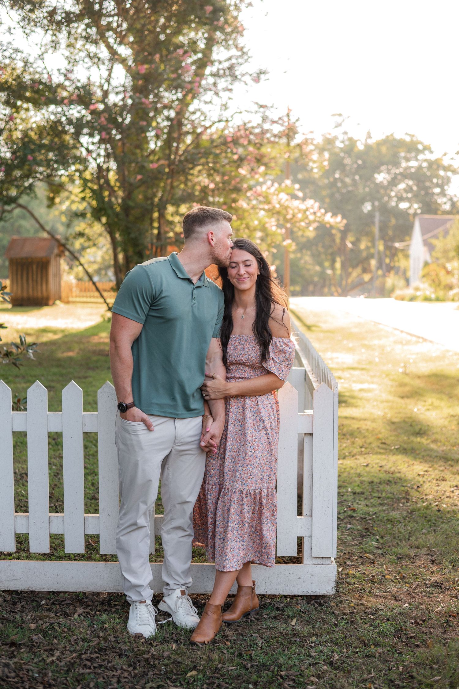 Romantic couple sharing a tender moment by a white picket fence in a sunny garden.