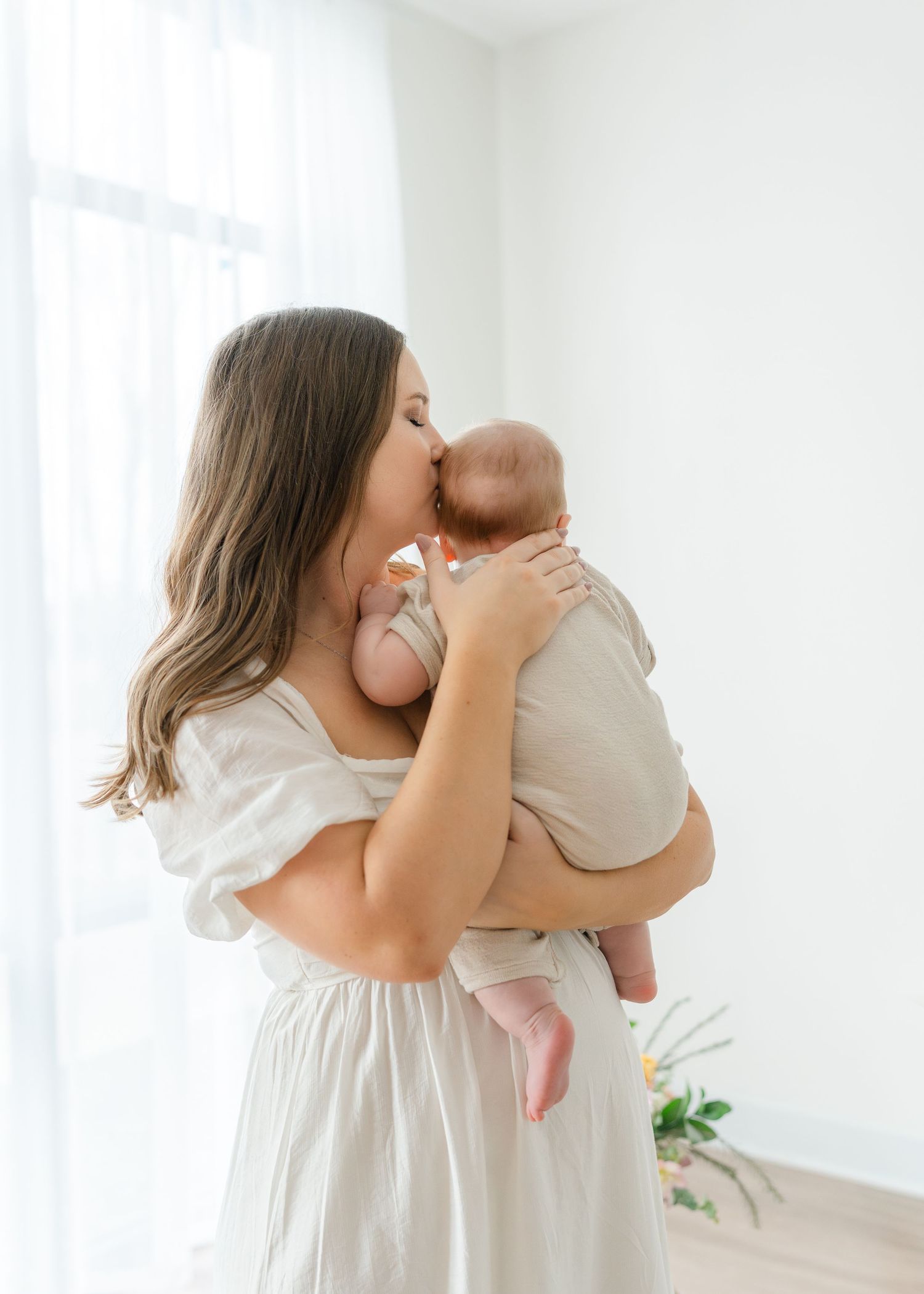 A mother in a white dress tenderly kisses her newborn baby while standing near a bright window.
