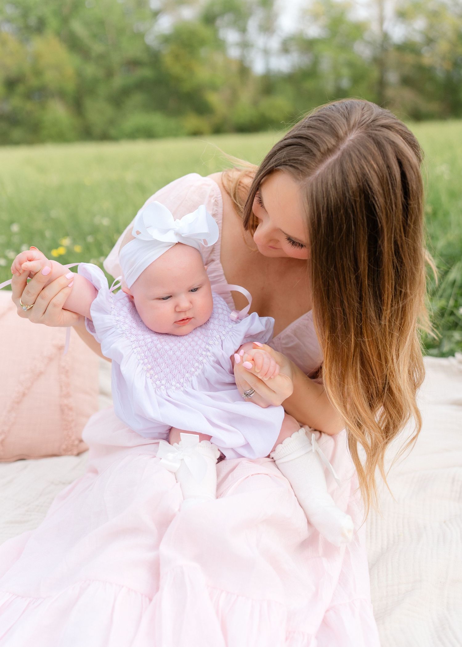 A mother in a pink dress holds her baby girl in a white bow while sitting on a blanket outdoors.